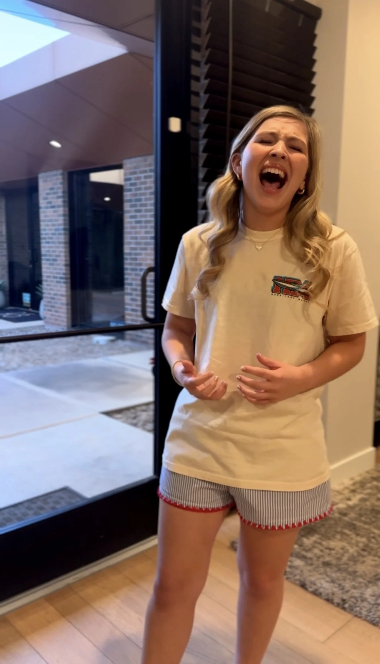 A young woman with long blonde hair, wearing a beige T-shirt with a logo and striped shorts with red trim, is standing indoors near a glass door with her mouth open and eyes closed, appearing to be singing or crying.