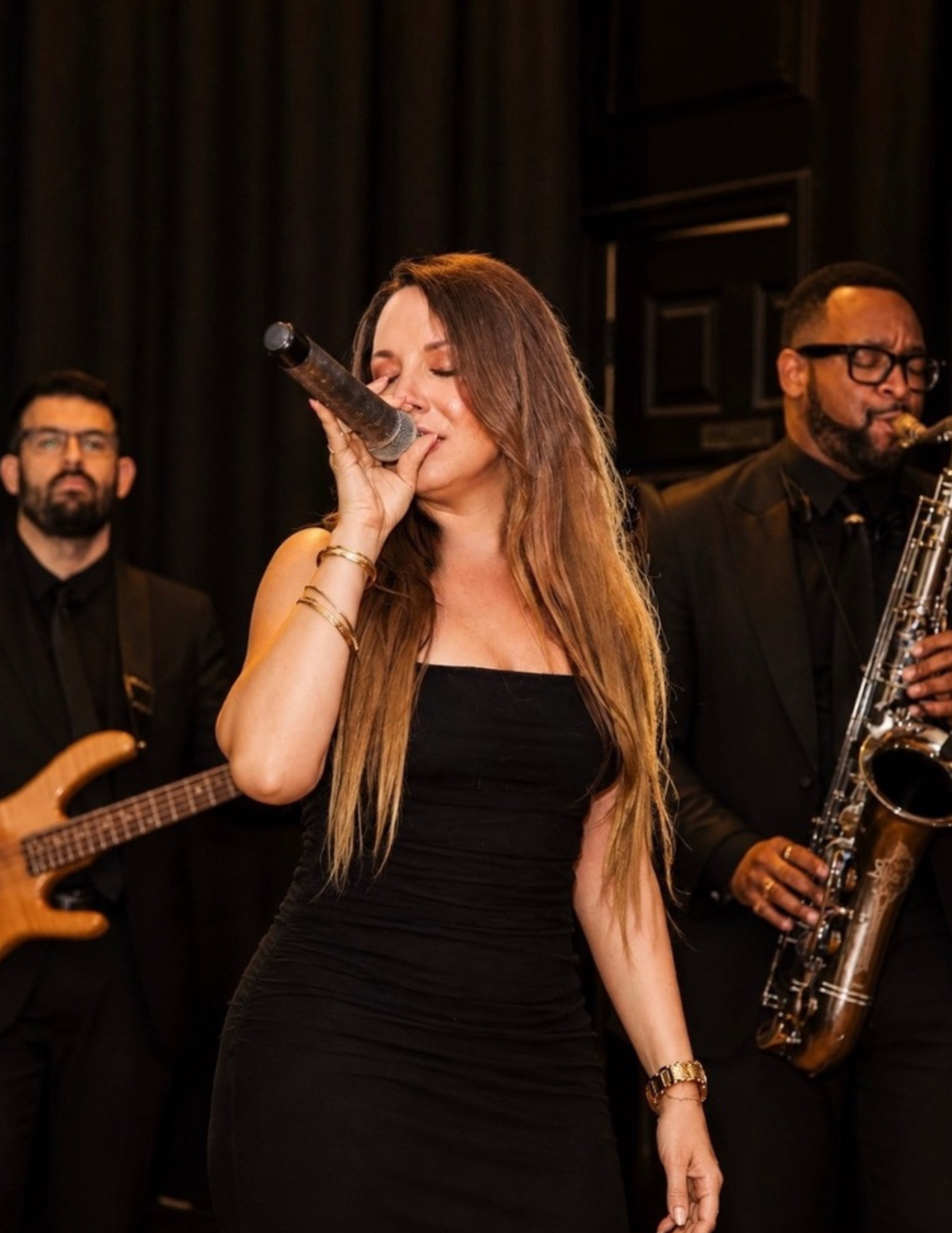 A woman in a black dress performing with a band on stage. She is singing into a microphone with her eyes closed. In the background, two men are playing instruments, one with a guitar and the other with a saxophone.