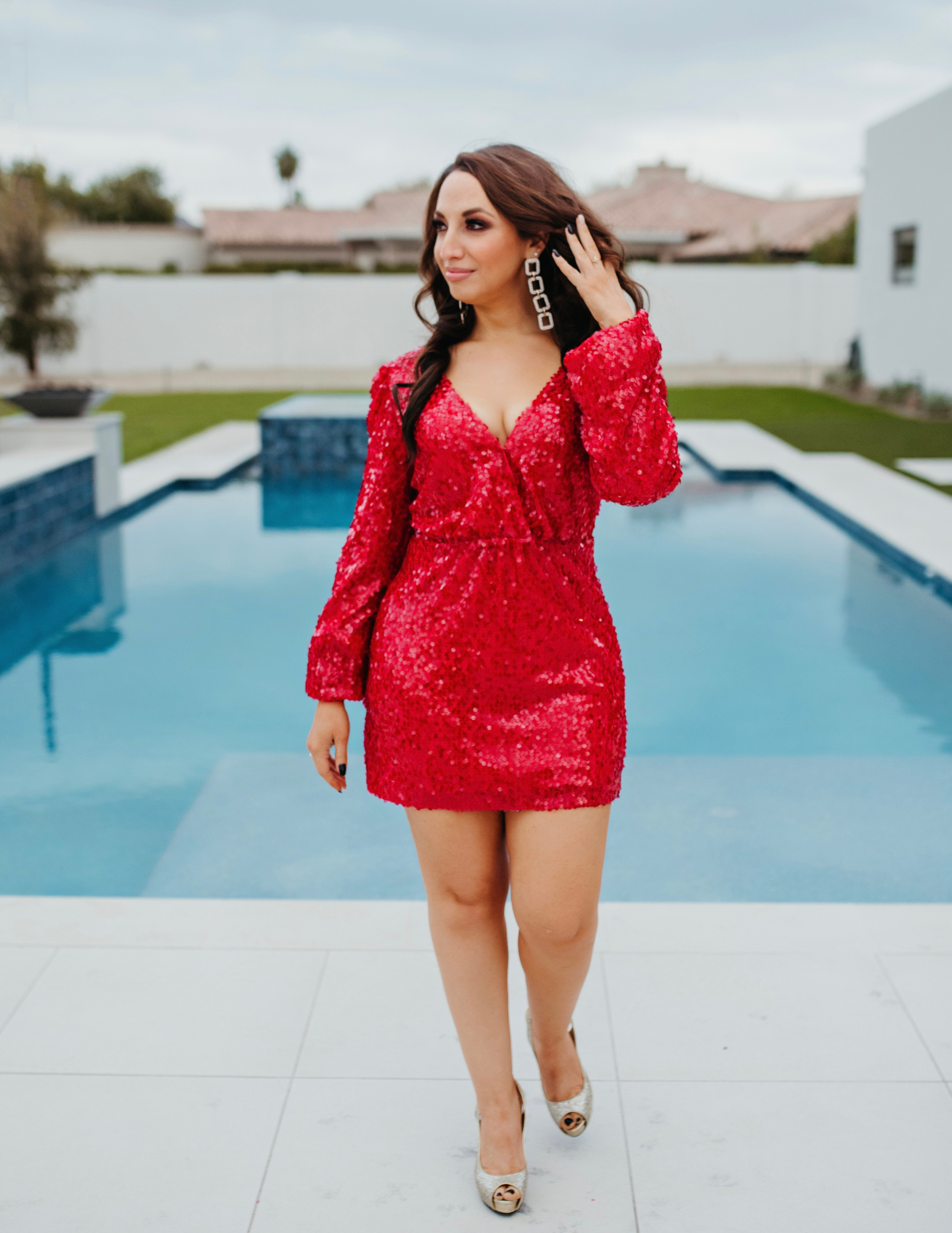 A woman in a red sequined dress with long dark hair walks past a swimming pool on a patio, wearing silver high heels and large geometric earrings.