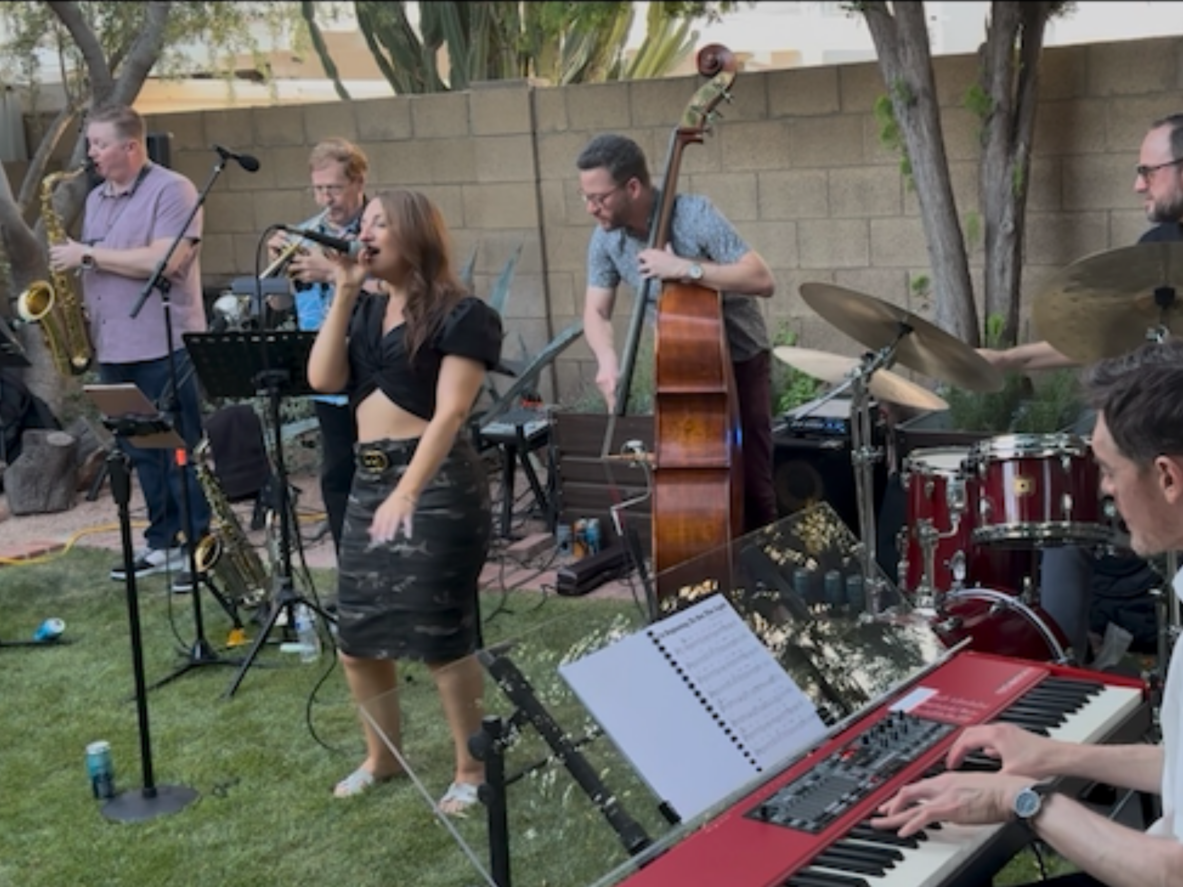 A group of musicians performing outdoors, including a woman singing into a microphone, a man playing a double bass, a pianist playing a keyboard, and others playing various instruments, with trees and a brick wall in the background.