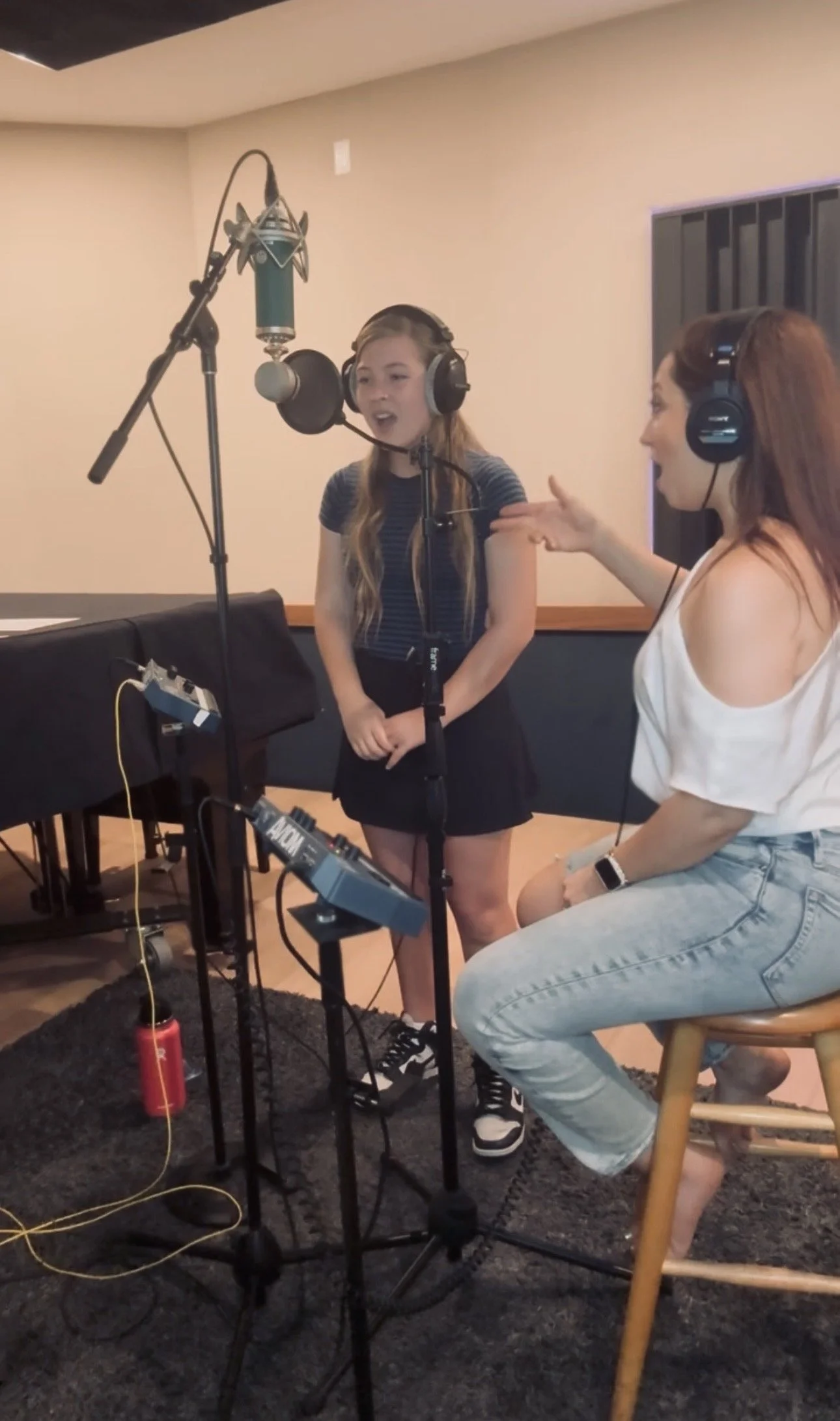 A young girl singing into a microphone in a recording studio with an adult woman coaching her. The girl is wearing a striped shirt, black skirt, and sneakers, while the woman is wearing a white top and jeans, seated on a wooden stool. There are audio recording equipment and microphones around them.