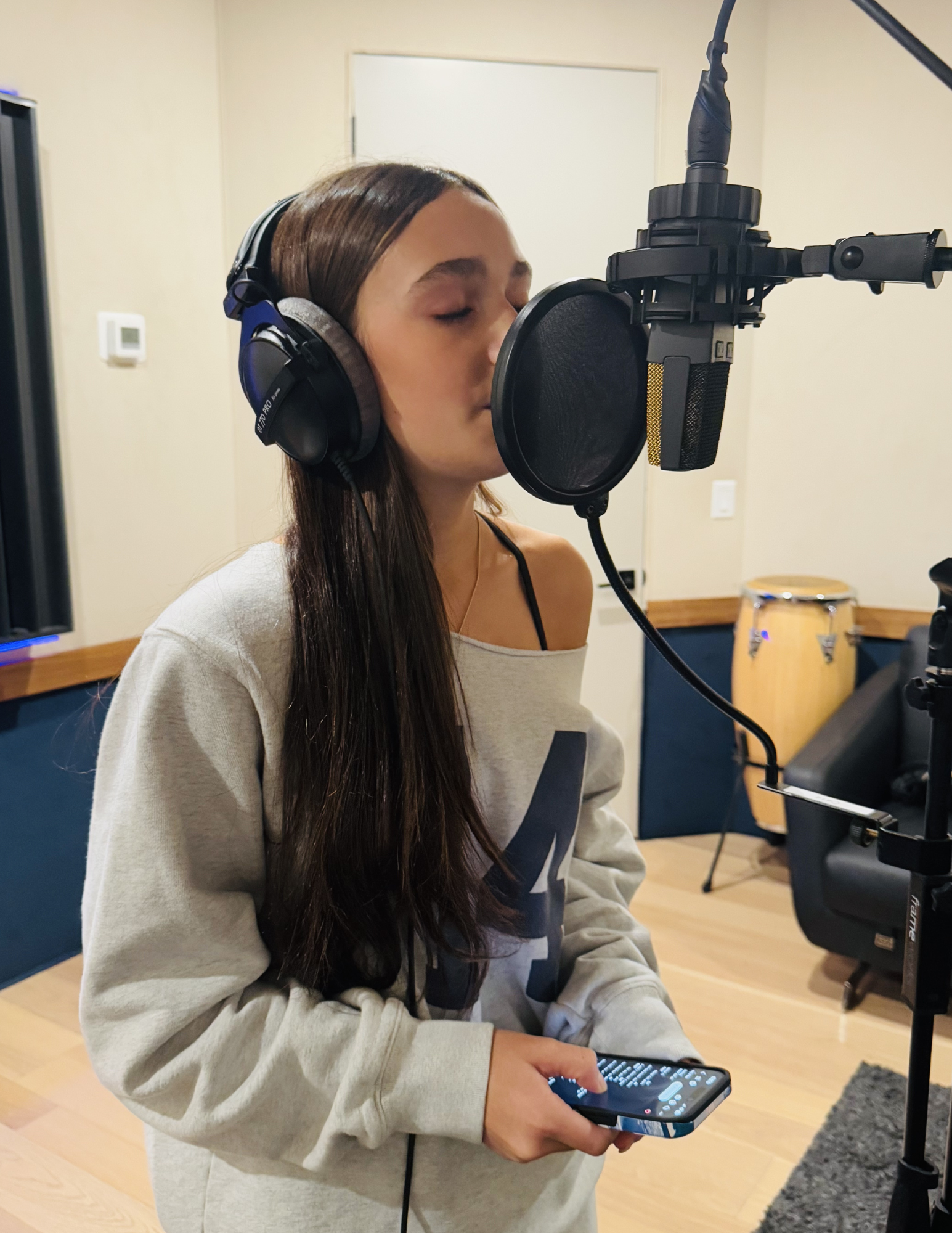 A young woman with long brown hair wearing headphones, standing in a recording studio, singing into a microphone with a pop filter, holding a smartphone in her left hand.