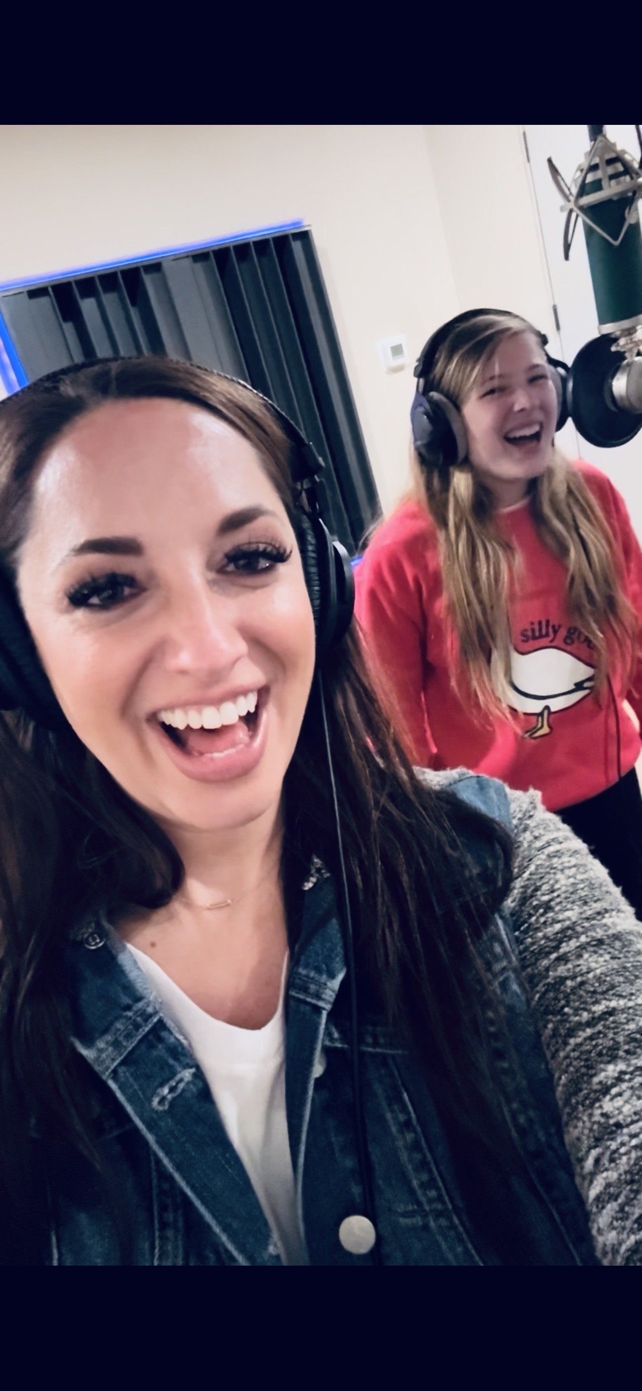 Two women wearing headphones in a recording studio, smiling and laughing at the camera.