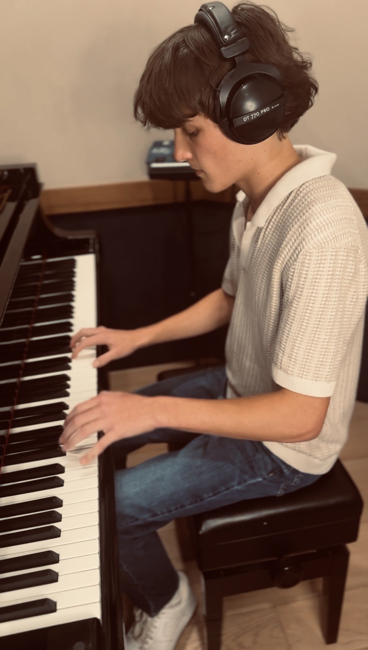 A young man wearing headphones playing a piano. He is sitting on a wooden chair, wearing a beige shirt and jeans, with a focused expression.