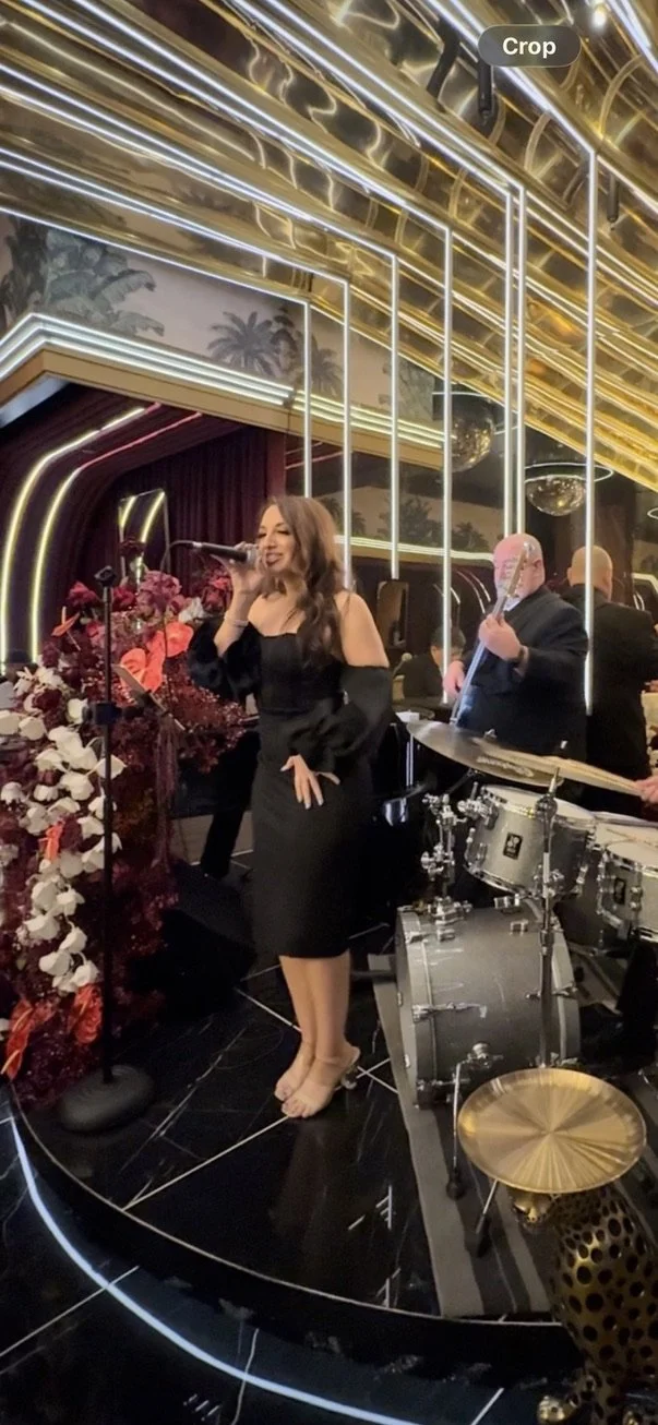 A woman in a black dress singing into a microphone on a stage with a band, surrounded by floral arrangements and elaborate lighting, in an upscale venue.