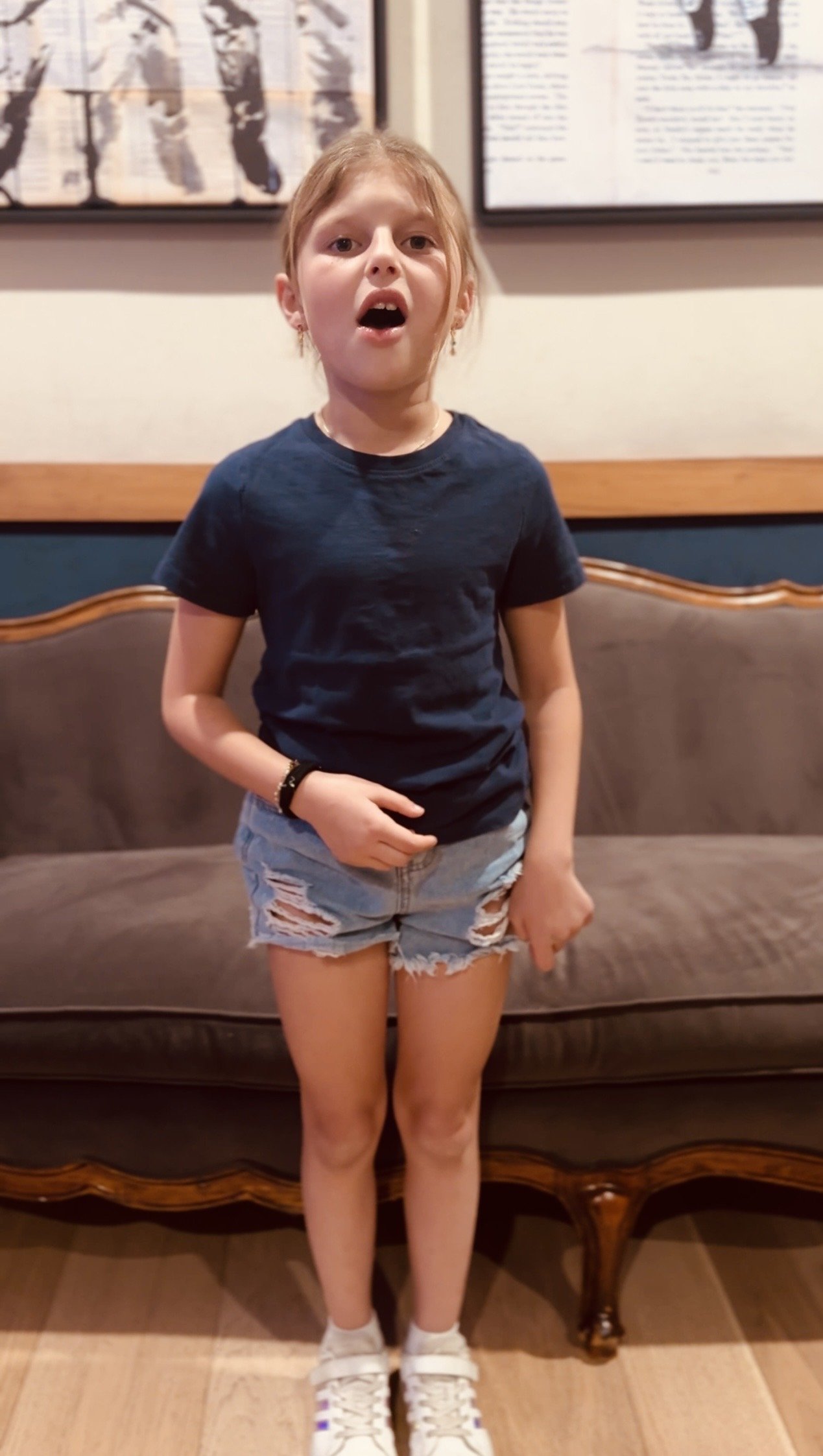 A young girl with long red hair wearing a dark t-shirt, ripped denim shorts, white sneakers, and a black wristband, standing in front of a brown couch with wooden trim, with framed photographs and artwork on the wall behind her.