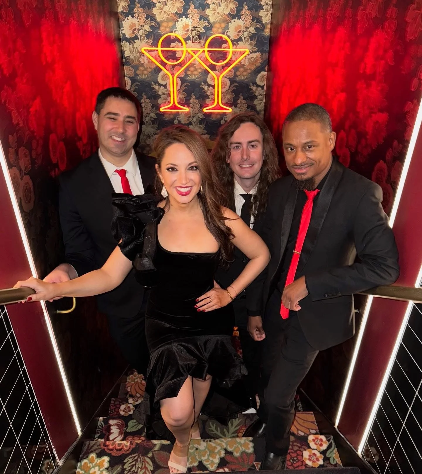 Four people in formal attire posing in front of a red and floral background with neon drink glasses and eyes sign overhead.
