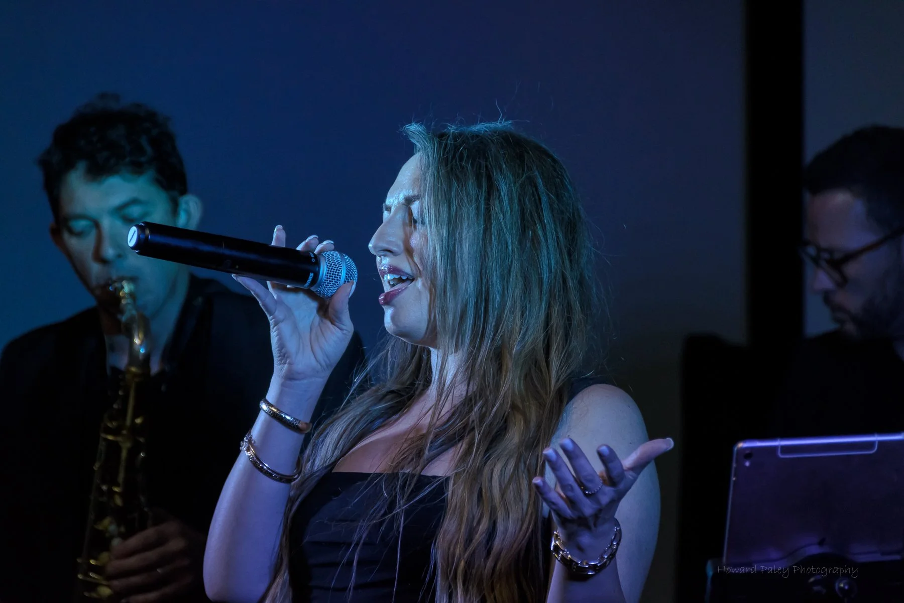 A woman singing into a microphone with two musicians in the background, one playing the saxophone and the other on a keyboard, in a dimly lit setting.