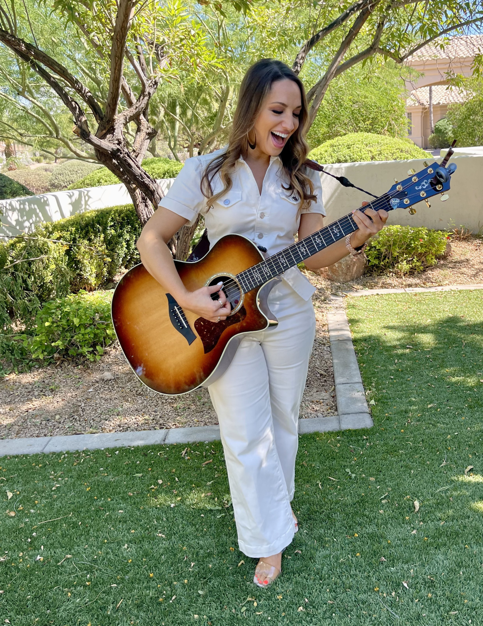 A woman smiling and playing an acoustic guitar outdoors in a garden with green trees and bushes.