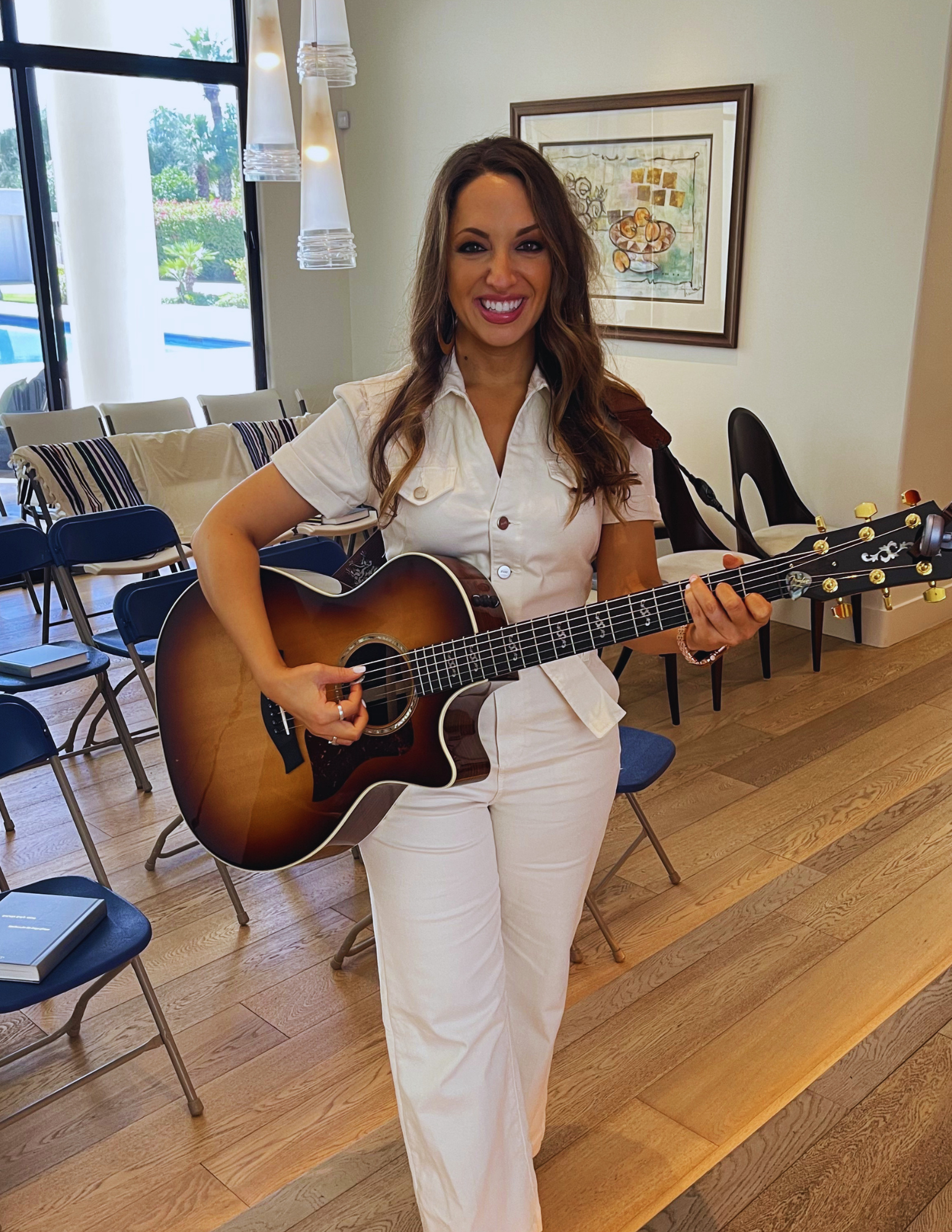 A woman with long brown hair wearing a white shirt and white pants smiling and playing an acoustic guitar in a room with wood flooring, chairs, a framed painting on the wall, and a large window showing outdoor greenery.