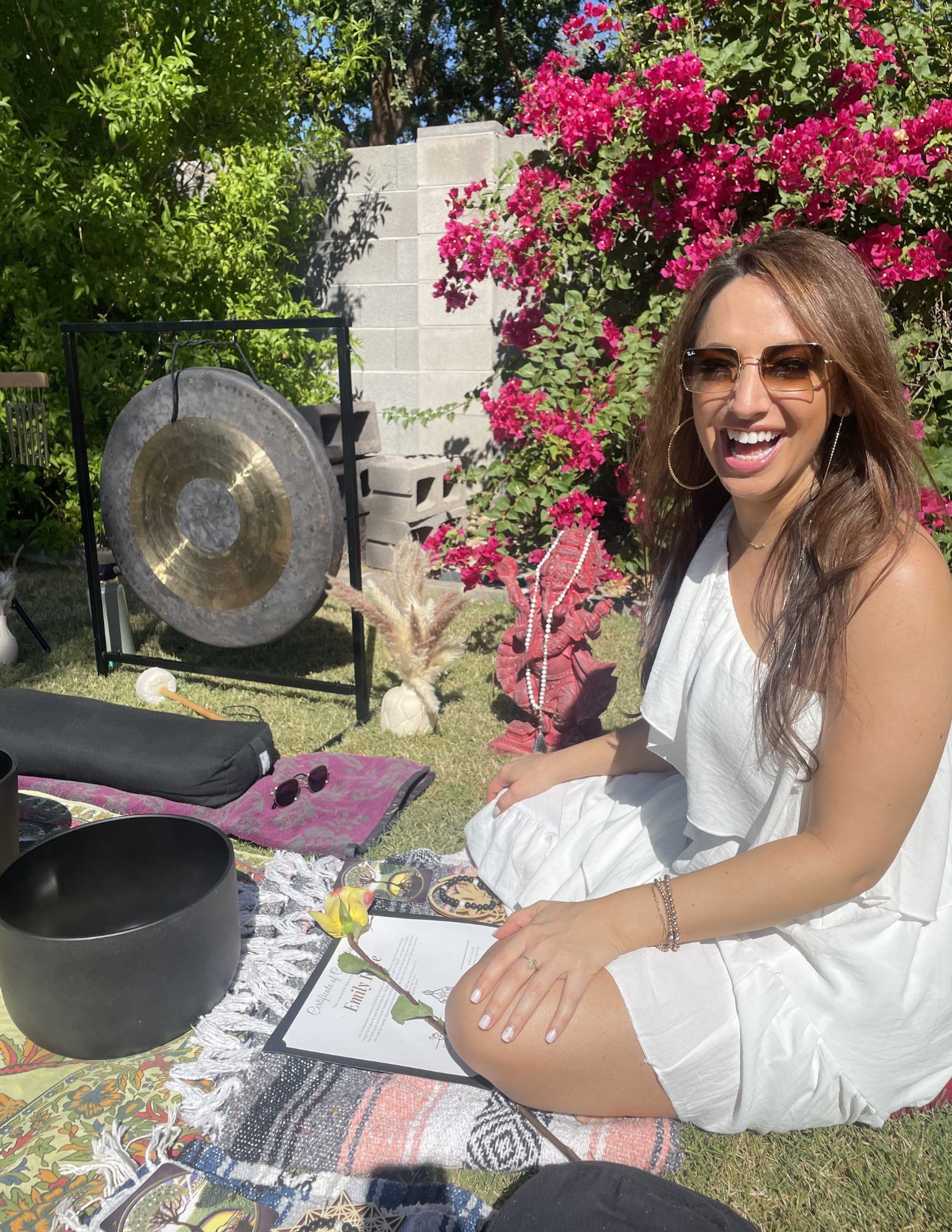 A woman with brown hair wearing sunglasses and a white sleeveless dress is smiling and sitting at an outdoor table with various objects, including a black singing bowl, a colorful cloth, gardening tools, and jewelry. There are pink flowering bushes, green foliage, and a cinder block wall in the background under a sunny sky.