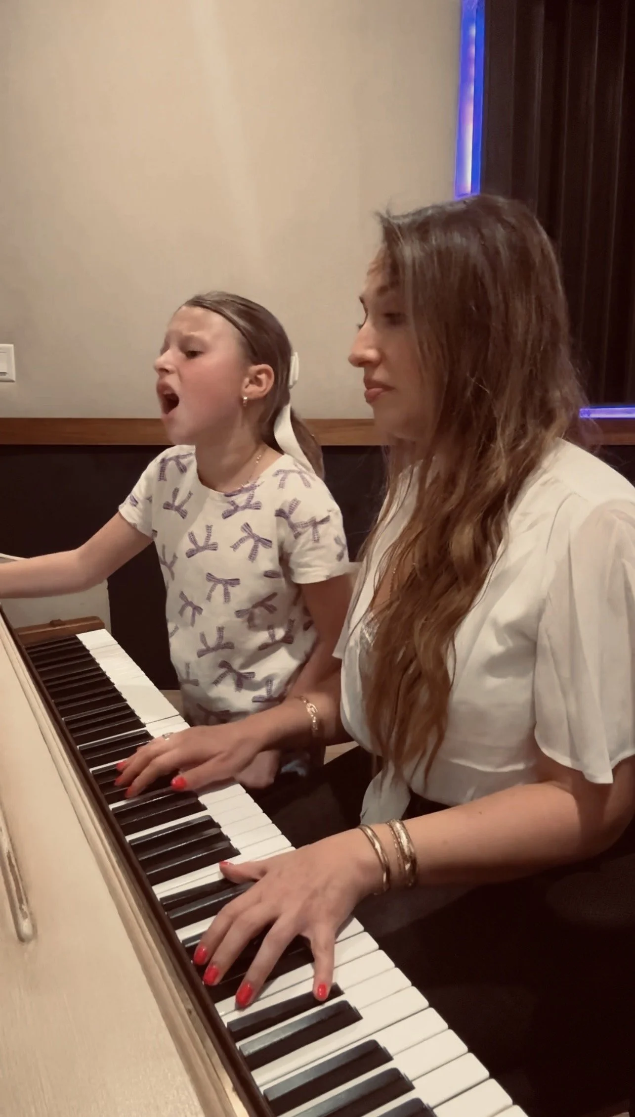 A woman with long red hair and a young girl with dark hair are sitting at a piano, playing and singing together. The woman is wearing a white shirt and several bracelets, while the girl is wearing a white t-shirt with purple bows. Both are focused on playing piano keys.
