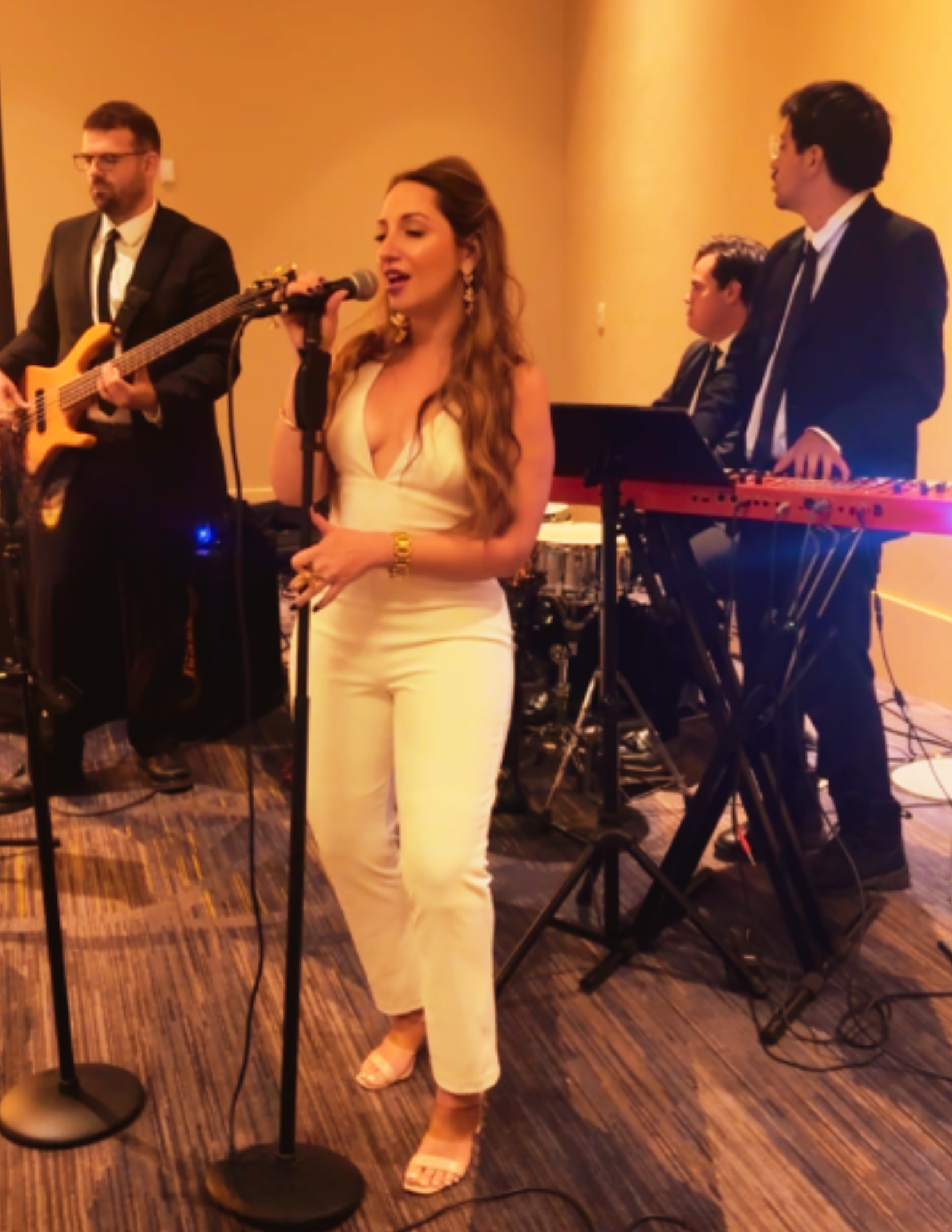 A woman singing into a microphone while four men, dressed in suits, play musical instruments in the background at an indoor event.