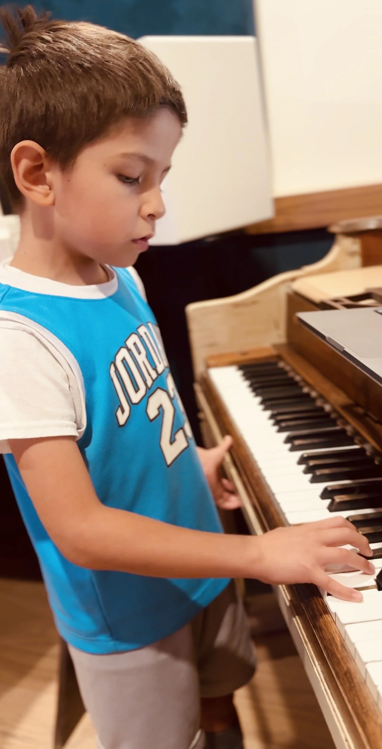 A young boy wearing a blue and white Jordan basketball shirt playing a wooden piano in a room.