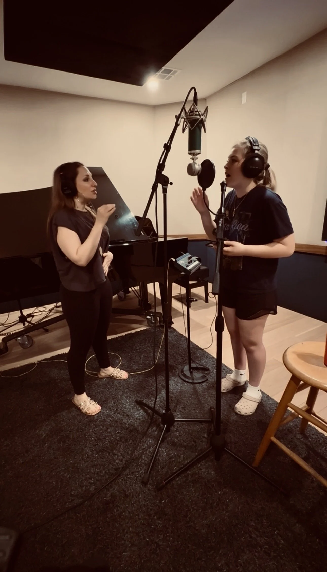 Two women are recording vocals in a music studio with a microphone and headphones. One woman is standing near a grand piano, wearing black clothes and floral slippers, while the other woman, also in casual clothes and slippers, is speaking into the microphone.