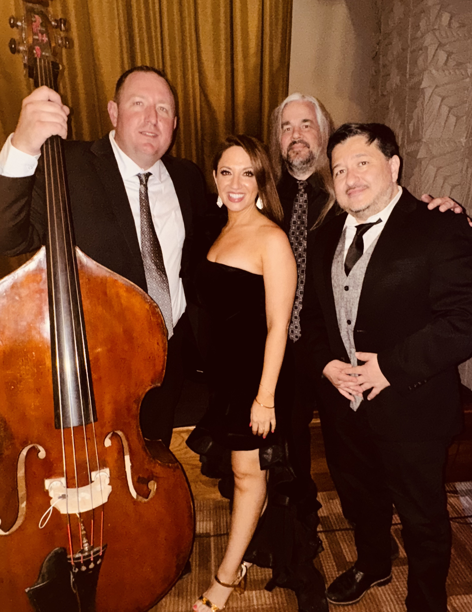Four people in formal attire pose together at an indoor event, with one person holding a double bass instrument visible on the left side of the image.