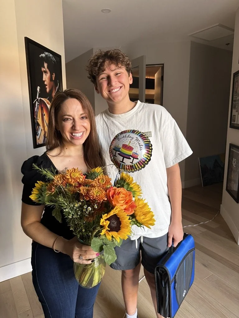 A smiling woman holding a bouquet of sunflowers and orange flowers next to a smiling teenage boy holding a birthday balloon and a backpack inside a home.
