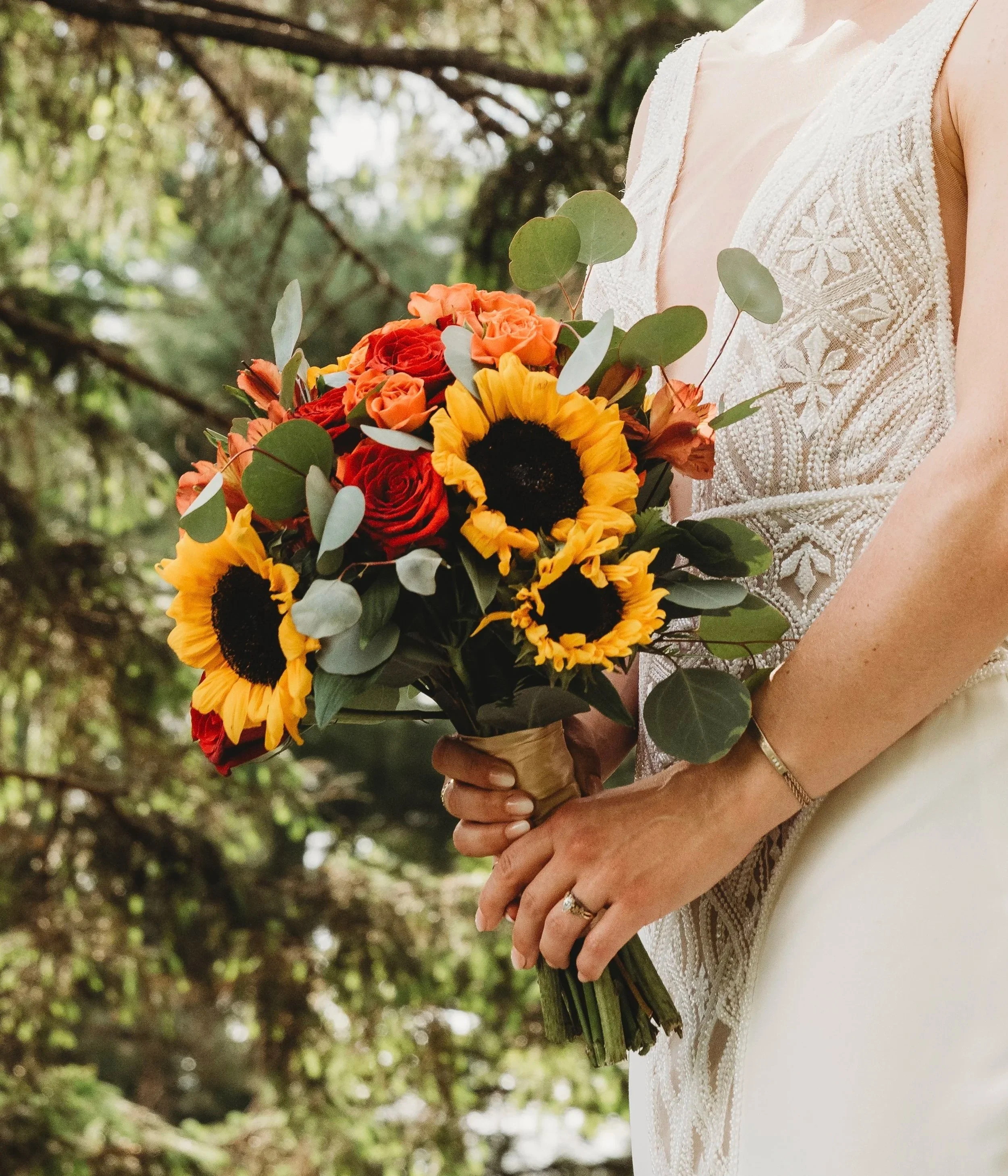A person holding a bouquet of colorful flowers, including sunflowers, roses, and other blooms, while wearing a white lace dress.