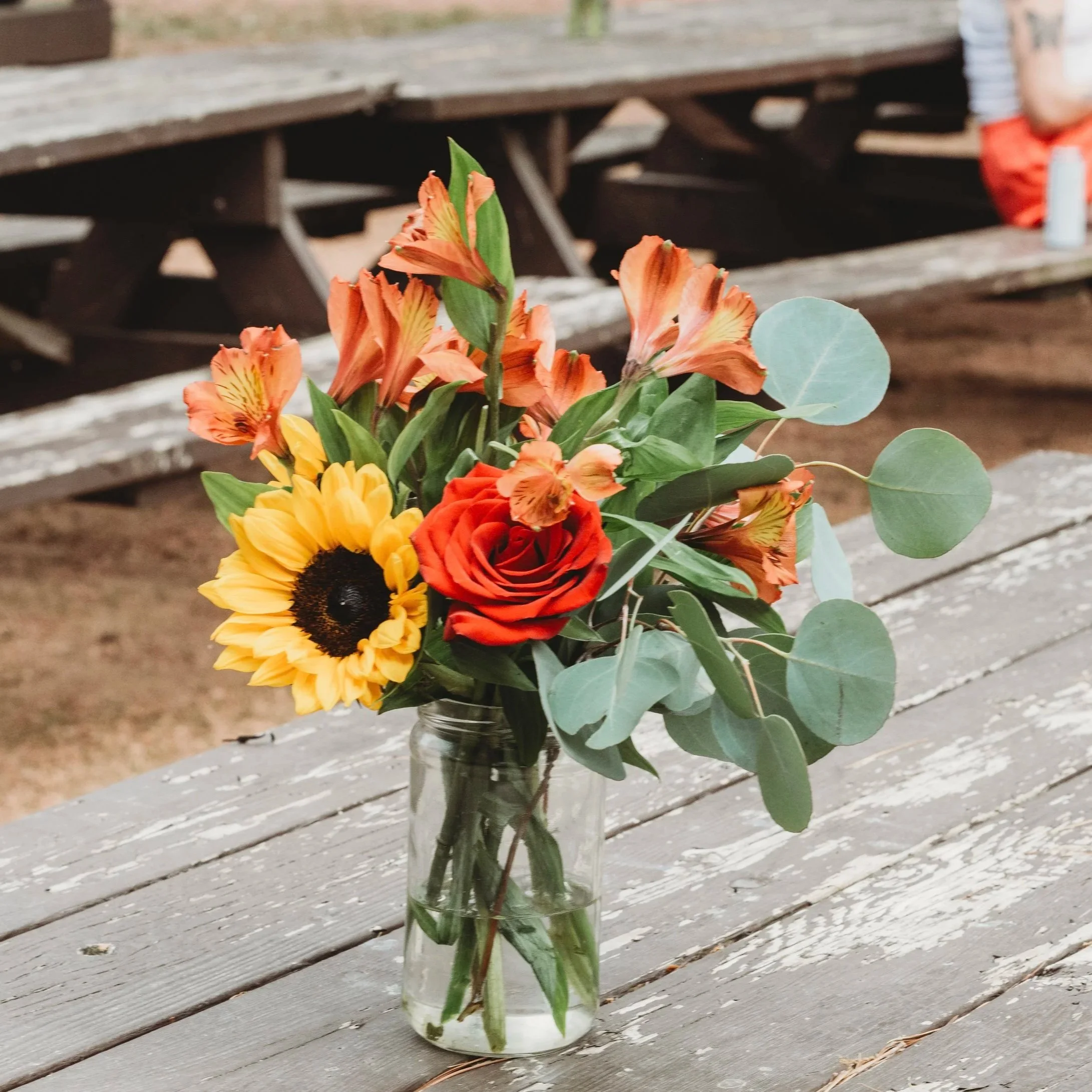 A glass jar holding a bouquet of orange, yellow, and red flowers with green leaves, placed on a weathered gray wooden surface.