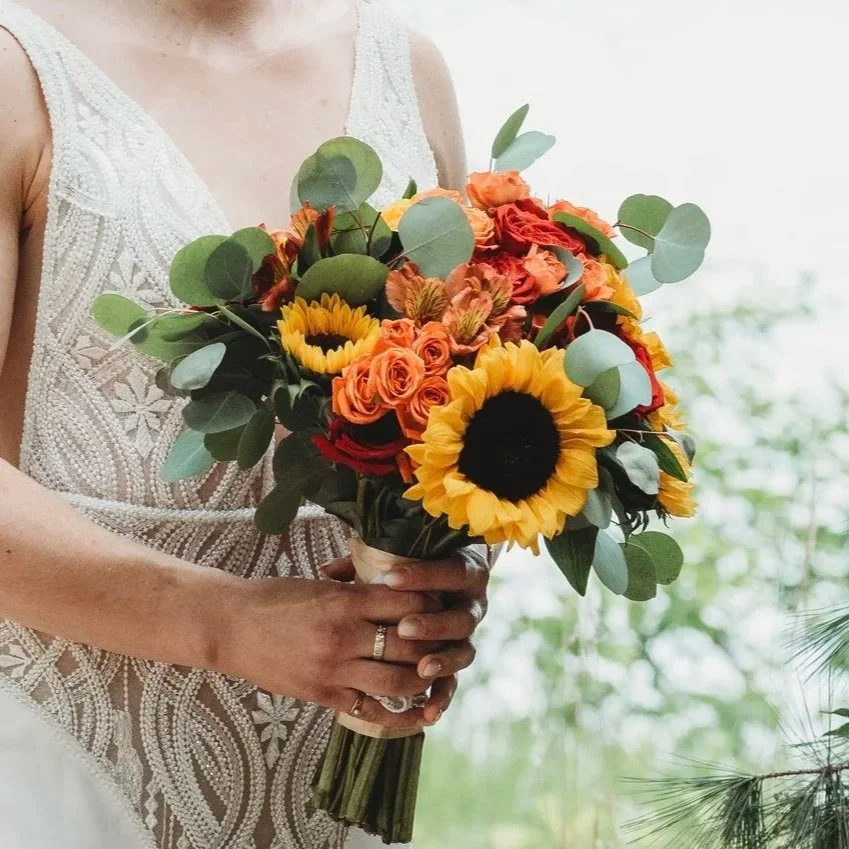 A woman in a white lace dress holding a bouquet of sunflowers, orange roses, and greenery.