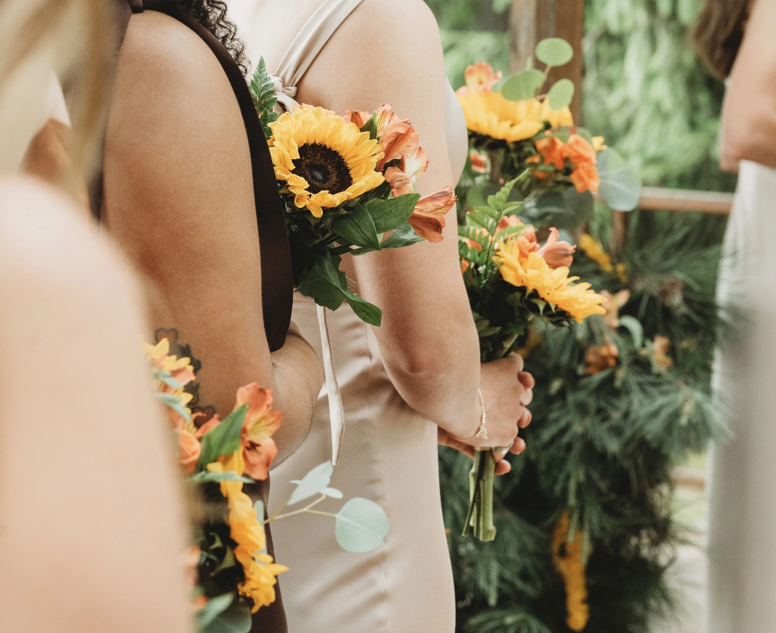 Group of women standing outdoors with floral bouquets, focusing on their torsos and arms, with a natural green background.