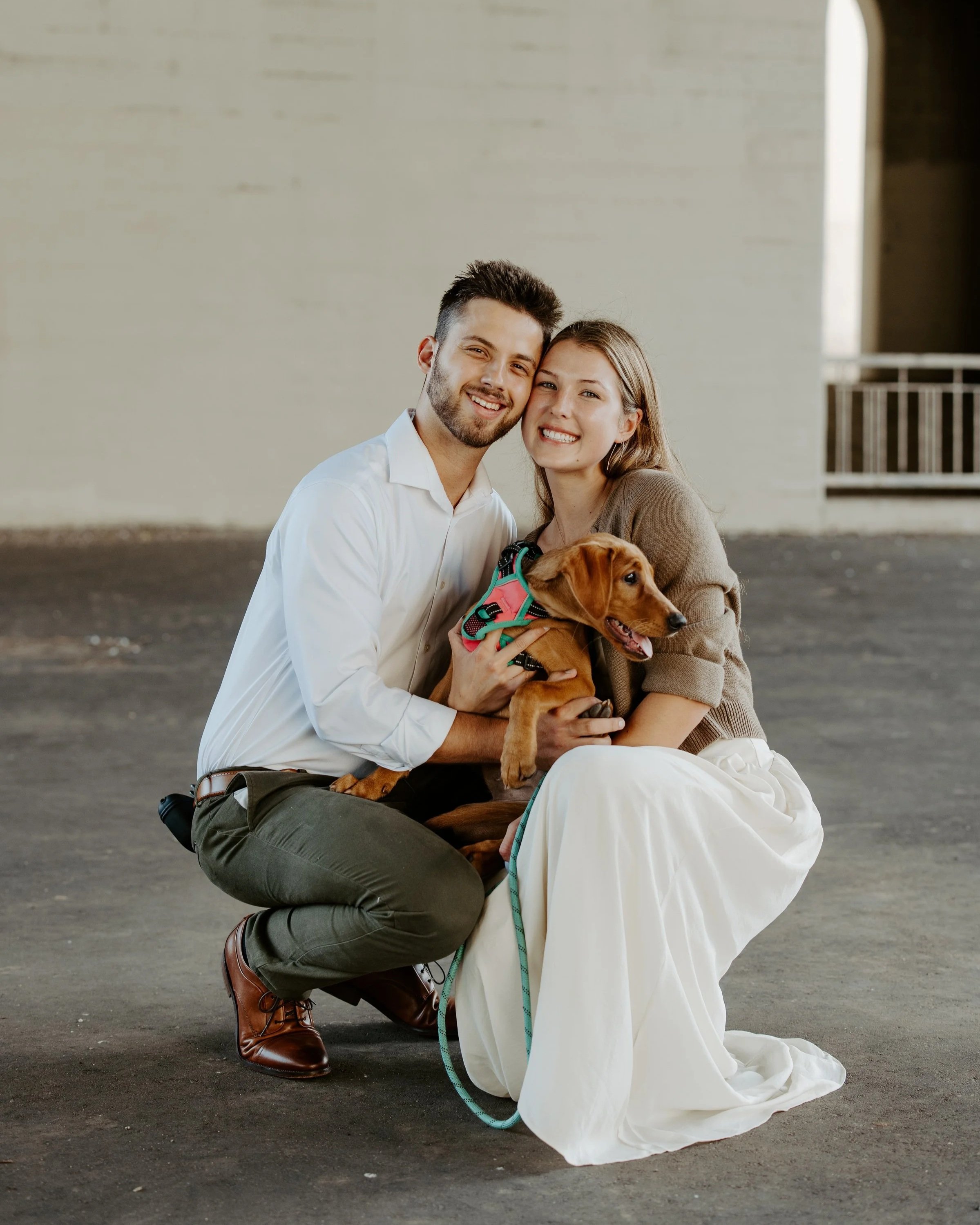 A man and a woman are sitting together on the ground, smiling, with a brown dog wearing a colorful harness between them. The man is wearing a white shirt and dark pants, and the woman is in a beige sweater and white skirt. The background is plain with an opening to the right.