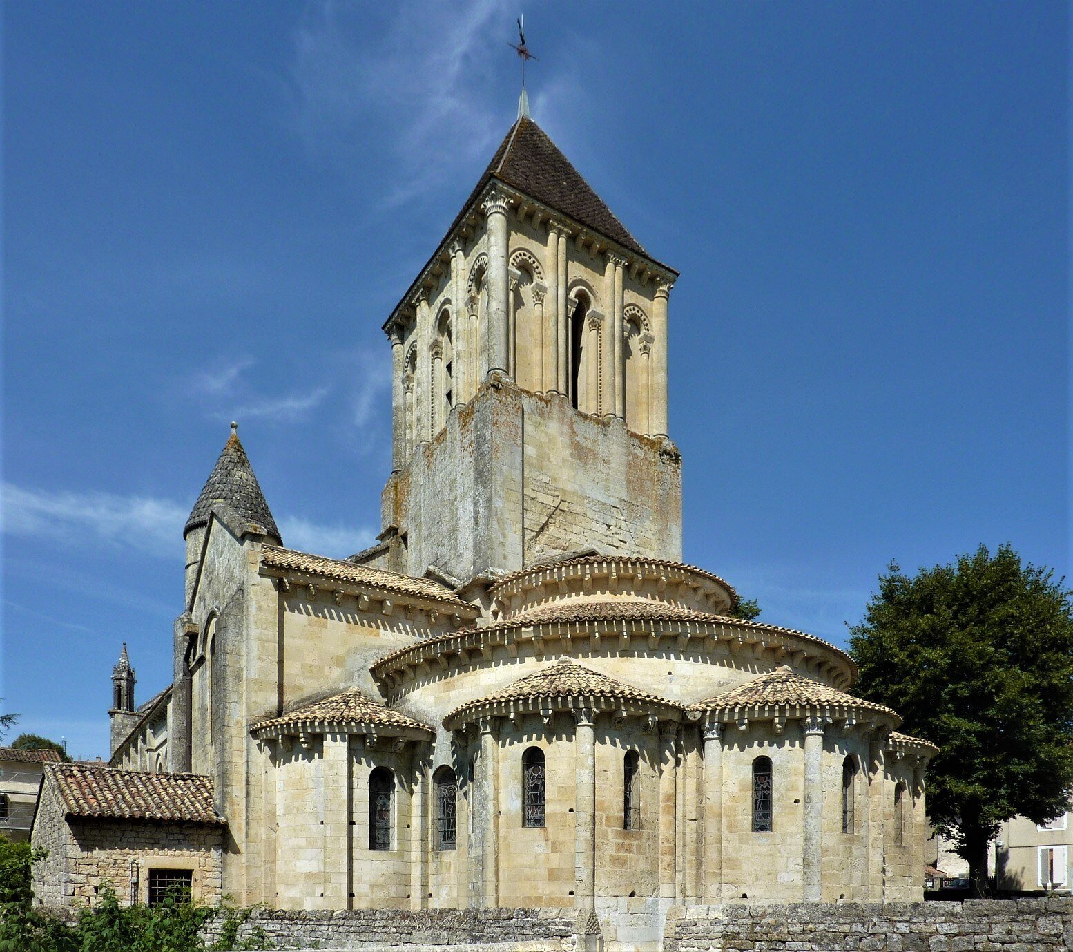 Église ancienne en pierre avec un clocher, toit en tuiles, et un hélicoptère volant au-dessus, sous un ciel bleu.