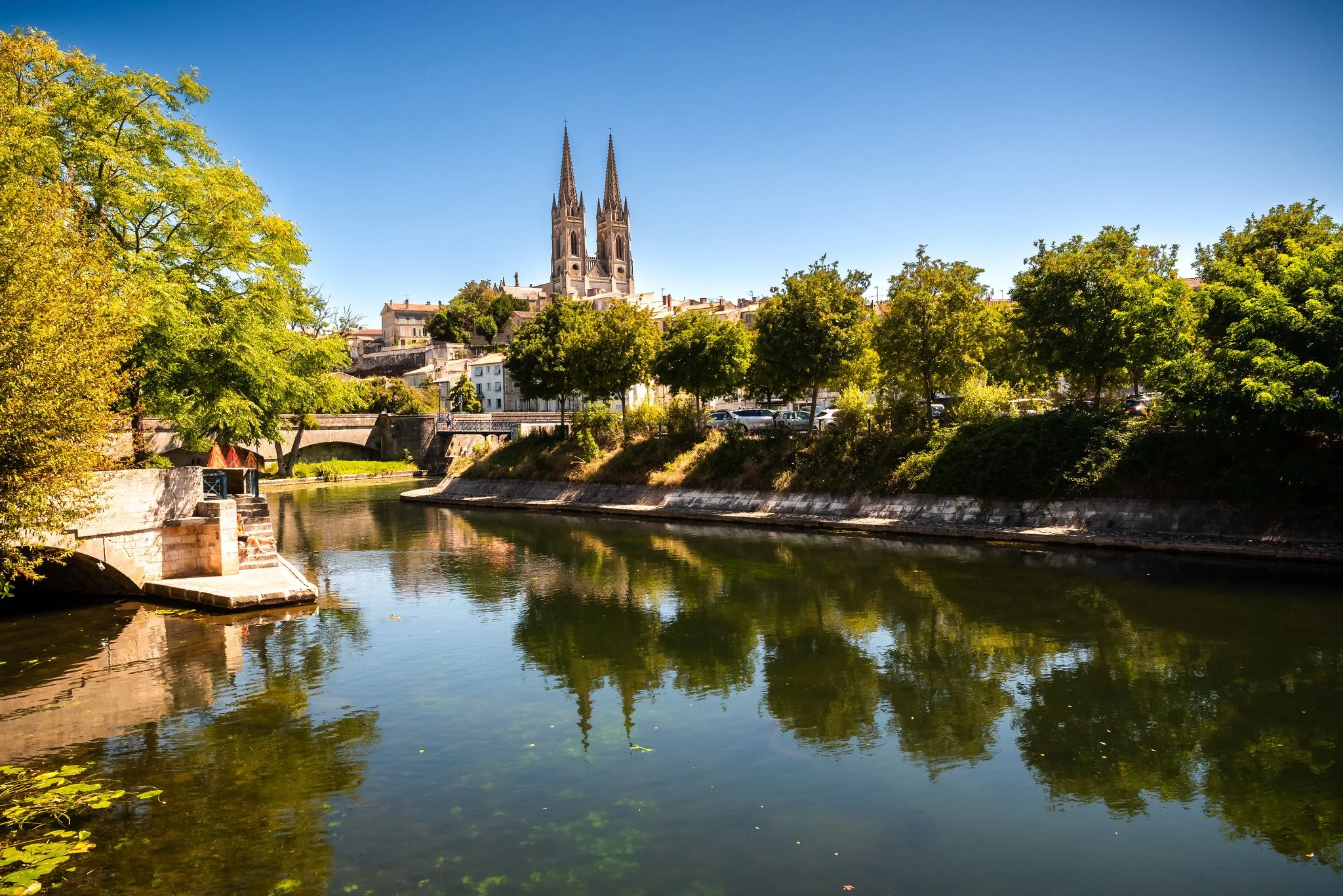 Une vue d'une rivière bordée d'arbres verts, avec en arrière-plan une ville et une grande église à deux flèches pointues sous un ciel bleu clair.