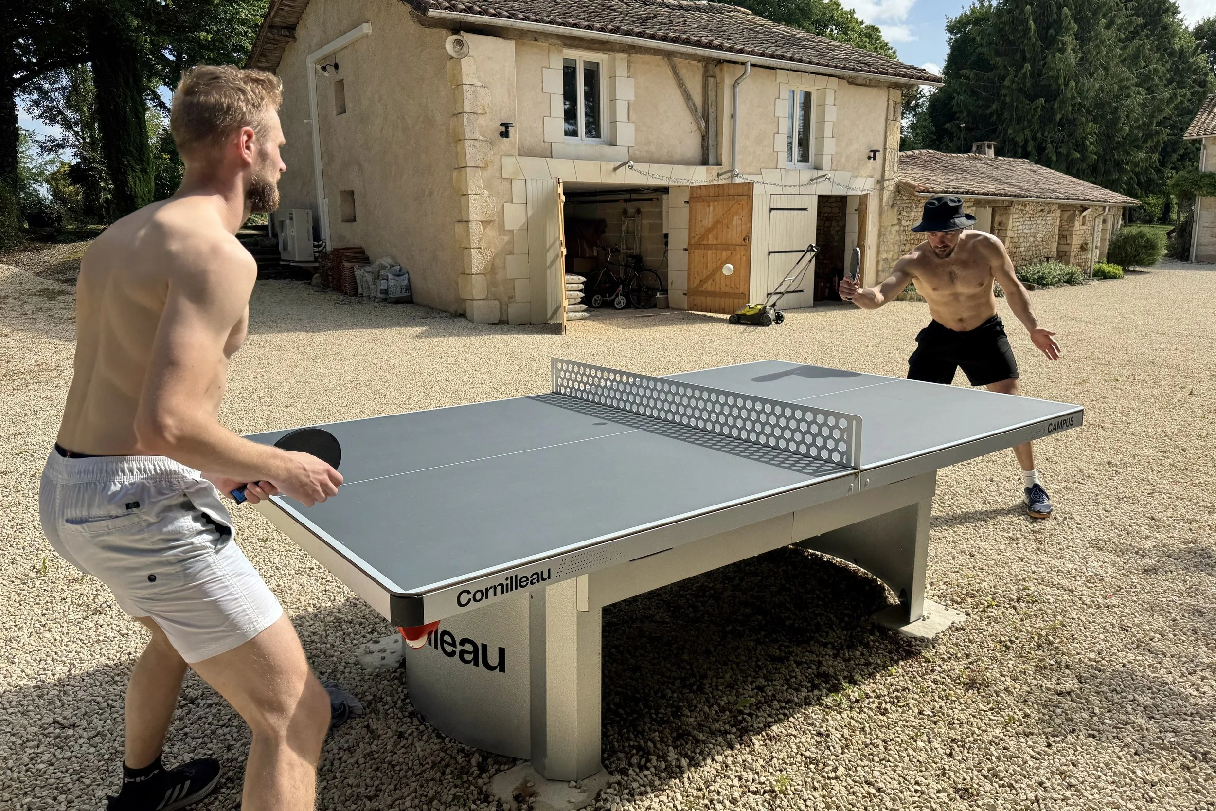 Deux hommes jouent au tennis de table dans une cour extérieure par une journée ensoleillée. Les deux sont en shorts, un avec un chapeau noir. La maison en pierre en arrière-plan a un garage ouvert avec des vélos et autres objets à l'intérieur.