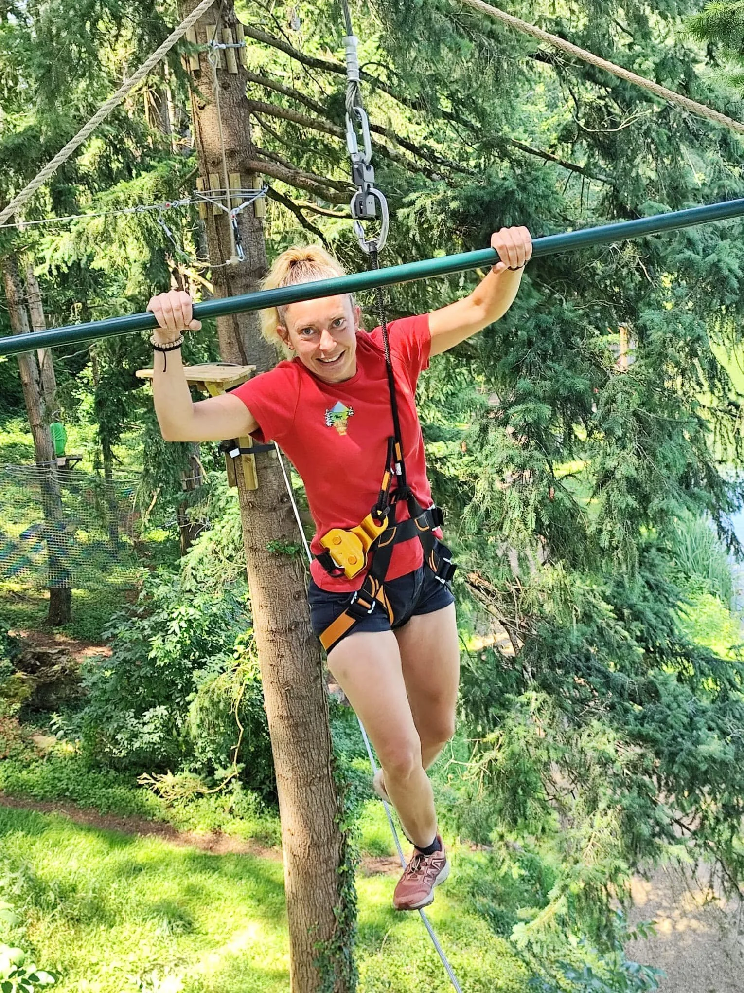 Une femme faisant de la tyrolienne dans une forêt verdoyante, portant un harnais de sécurité et un t-shirt rouge.