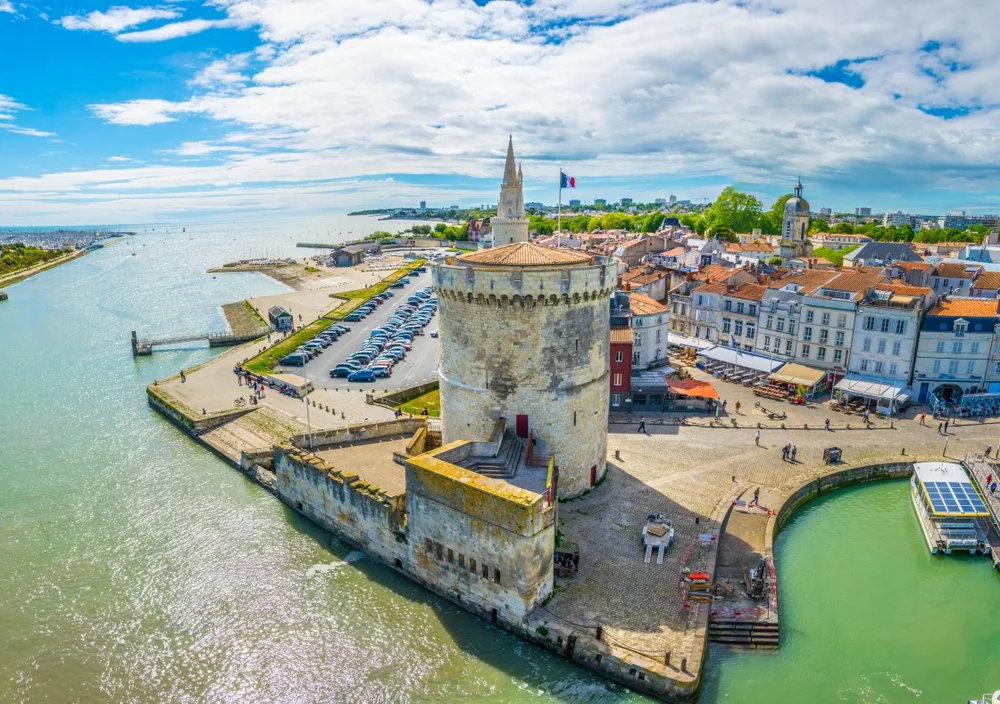 Vue aérienne d'une vieille forteresse avec un tour rond au premier plan, une tour avec un clocheau à l'arrière, et une place avec des bâtiments en pierre et des restaurants, entourée d'une rivière, sous un ciel partiellement nuageux.