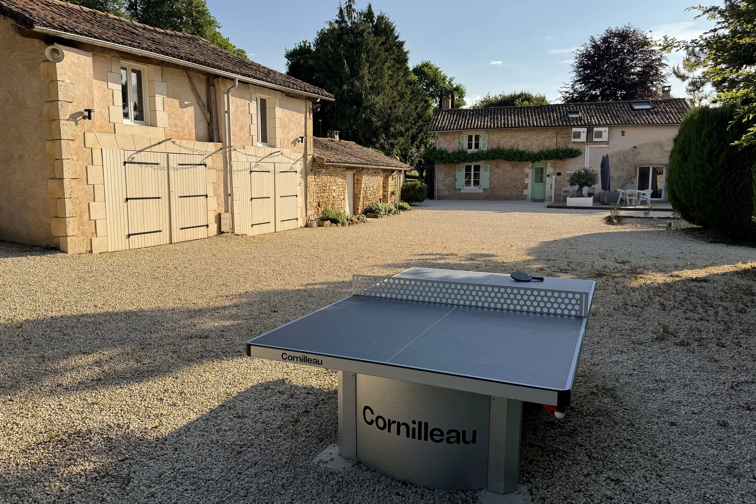 Une cour extérieure avec deux maisons en pierre, des arbres, un mobilier de jardin, et une table de ping-pong. L'ambiance est calme et ensoleillée.