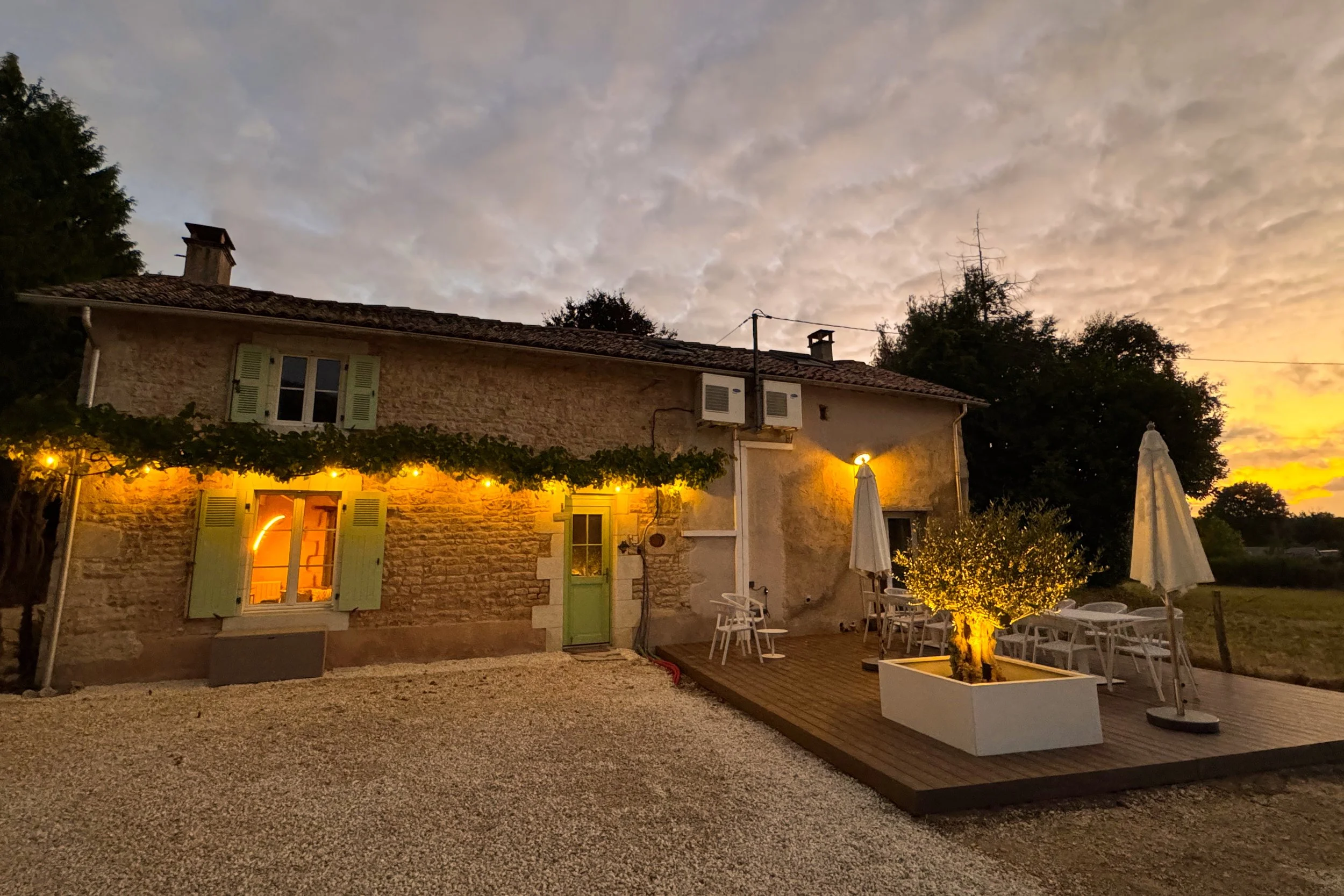 Une maison en pierre avec des volets verts, éclairée chaleureusement à l'intérieur, sous un ciel nuageux au coucher du soleil, avec une terrasse extérieure équipée de parasols et de mobilier, entourée de végétation.