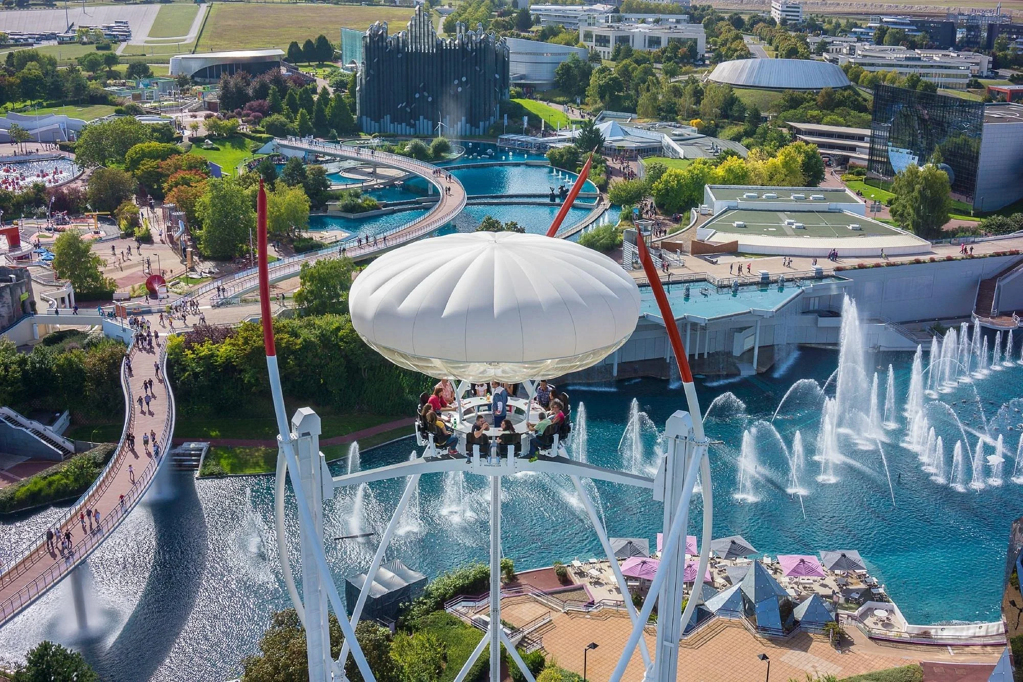 Vue aérienne du parc d'attractions Expo 2020 à Dubaï avec des attractions aquatiques, des jardins verts, un grand bâtiment d'inspiration futuriste, et une grande structure en forme de champignon blanche dans le premier plan.