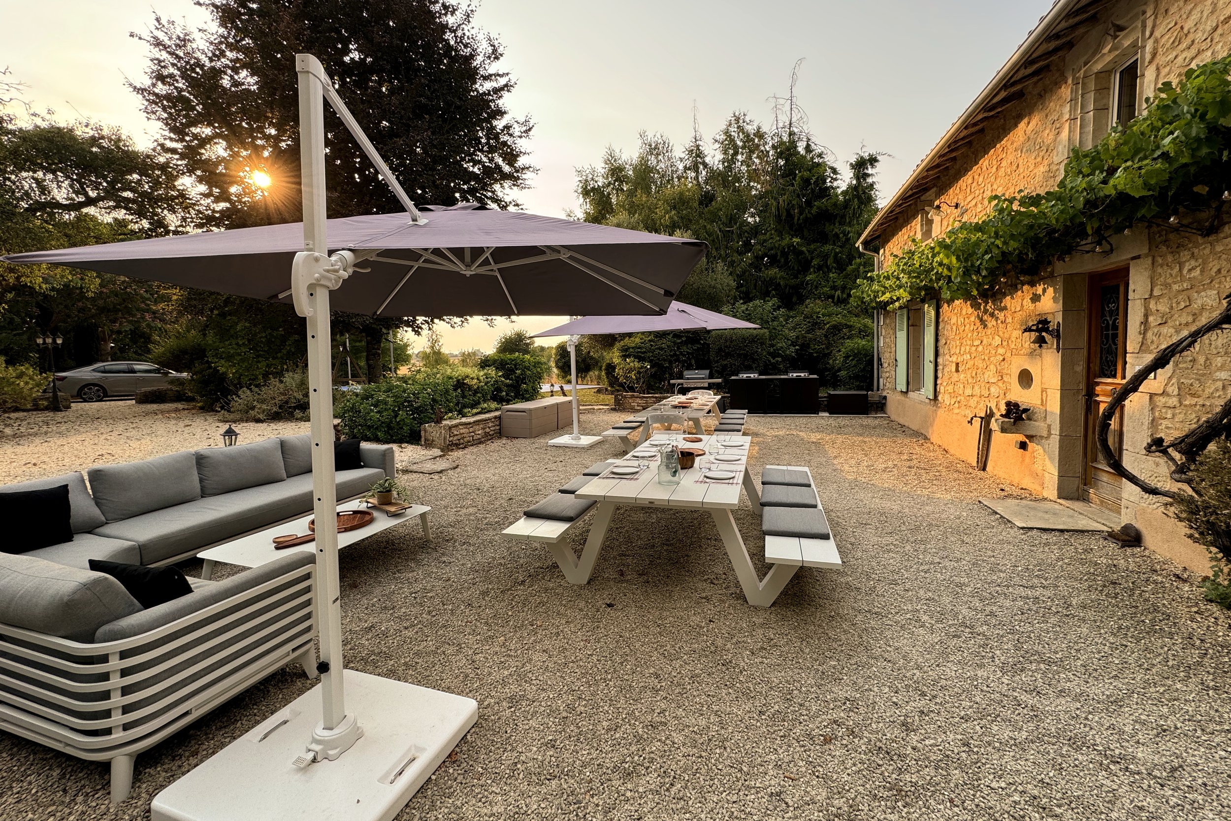 Terrasse extérieure avec une grande table blanche, plusieurs chaises, des parasols violets, un canapé gris, une maison en pierres, un barbecue, et un paysage avec des arbres en soirée.