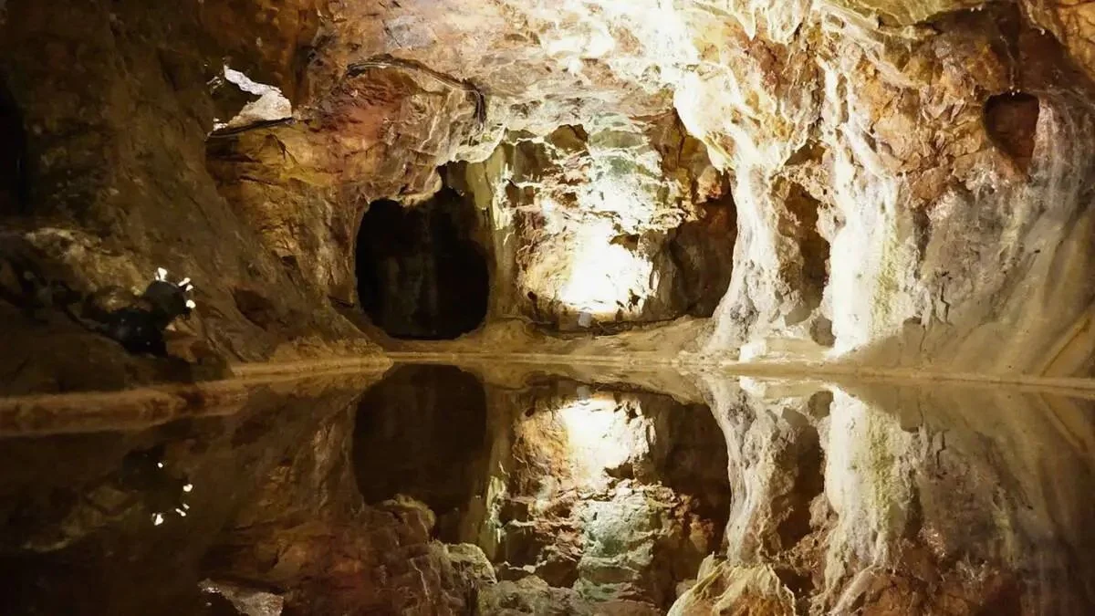 Caverne souterraine avec une grande ouverture à l'avant, des parois rocheuses éclairées, et un plan d'eau réfléchissant le plafond rocailleux.