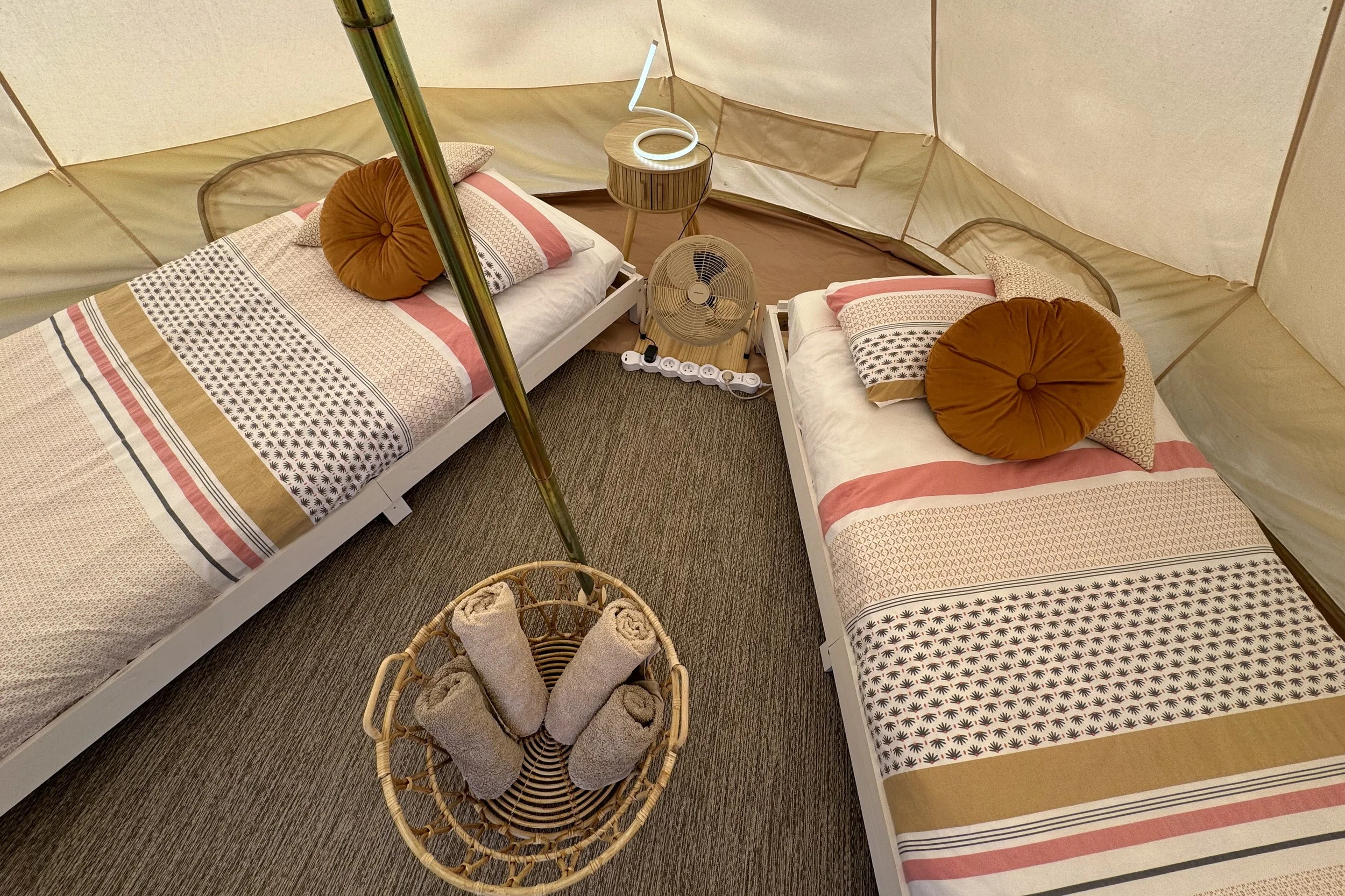 Deux lits simples avec draps et coussins ronds orange dans une tente. Entre les lits, une petite table en bois avec une lampe lumineuse en forme de spirale. En face, un ventilateur électrique et une panière en osier avec des serviettes roulées.