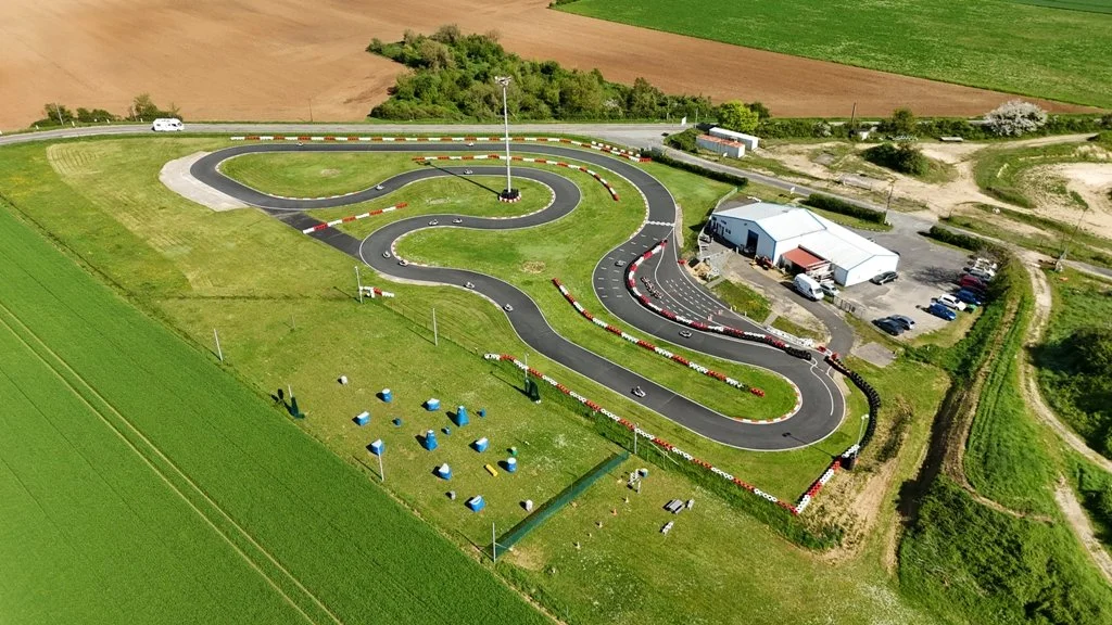 Piste de karting en extérieur avec des virages serrés, entourée de champs et de bâtiments, vue aérienne.