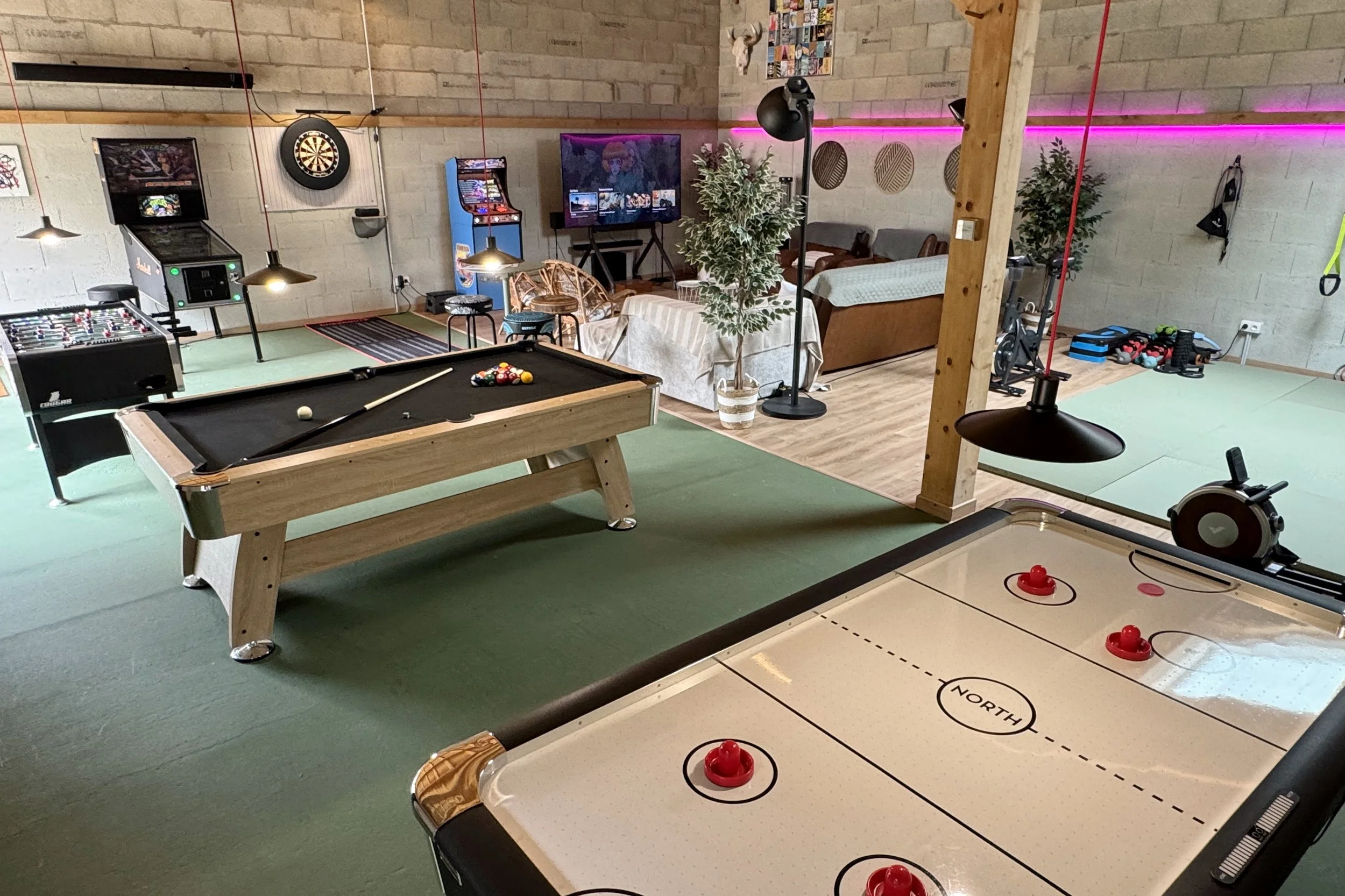 Une pièce de loisirs avec un billard, une table de hockey sur air, un baby-foot, et un coin détente avec des canapés et une télévision, décorée avec des plantes et un éclairage coloré.