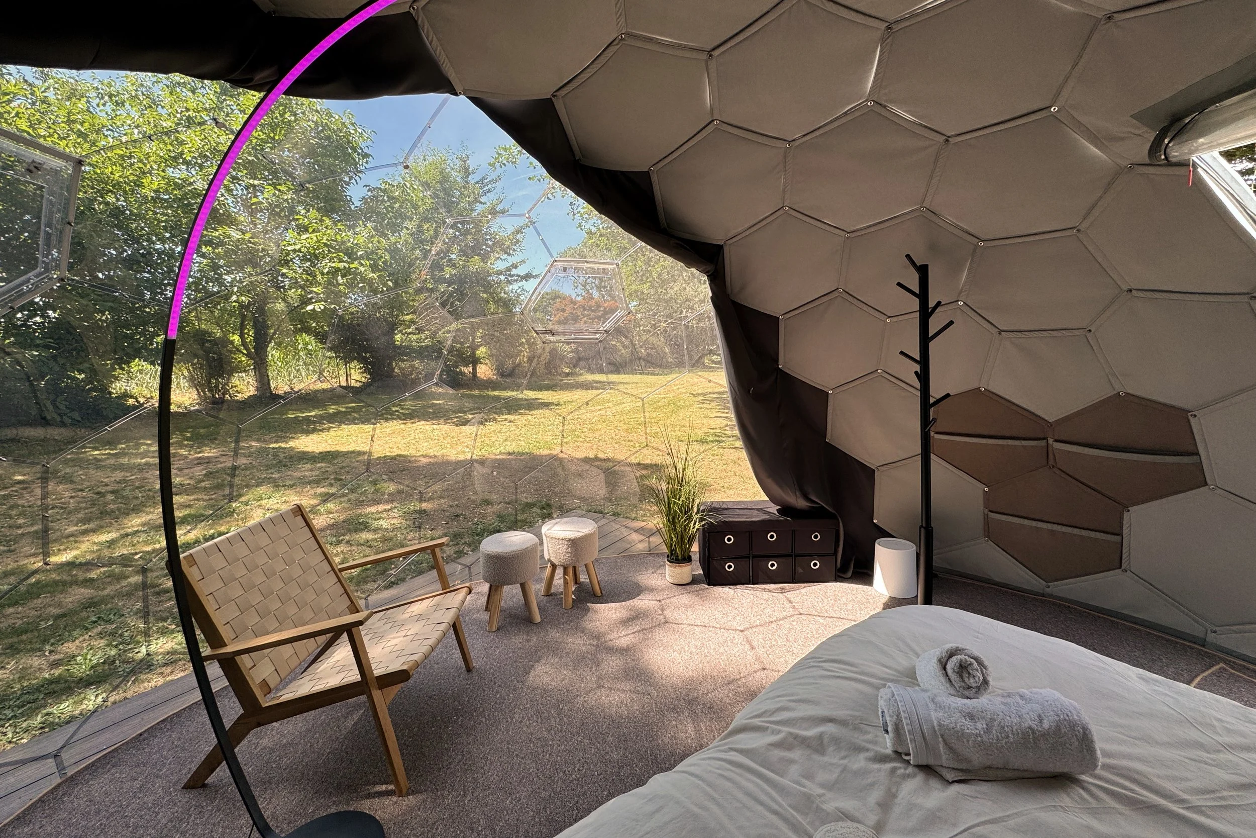 Vue de l'intérieur d'une structure en verre avec un plafond en forme d'échelle hexagonale. À l'extérieur, on voit des arbres et un ciel bleu. À l'intérieur, il y a un lit avec des serviettes roulées, une chaise en bois, deux petits tabourets, une plante en pot, une table de rangement noire, une lampe sur le sol, et des coussins.