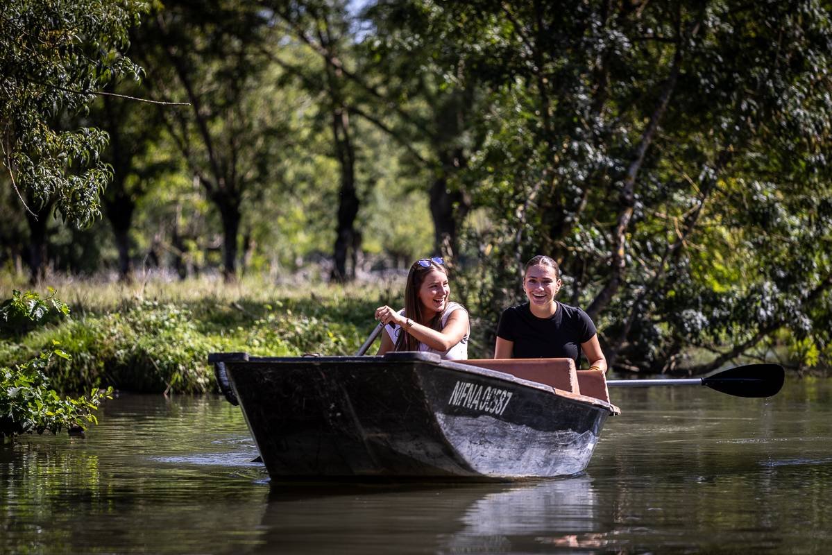 Deux jeunes femmes rient en pagayant dans un petit bateau sur un fleuve entouré d'arbres verts.