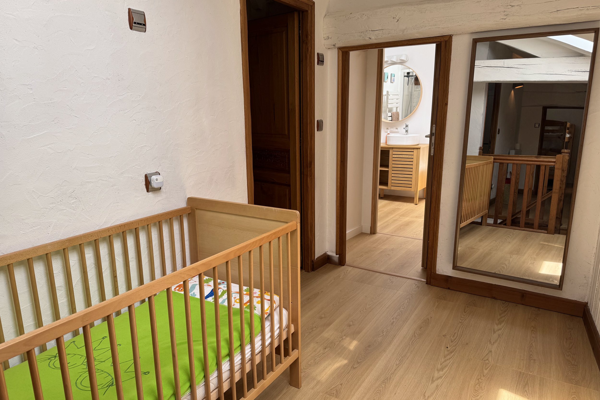 Une vue intérieure d'une pièce avec un lit bébé en bois, un mur blanc, un miroir rond, une salle de bain attenante, et des surfaces en bois.