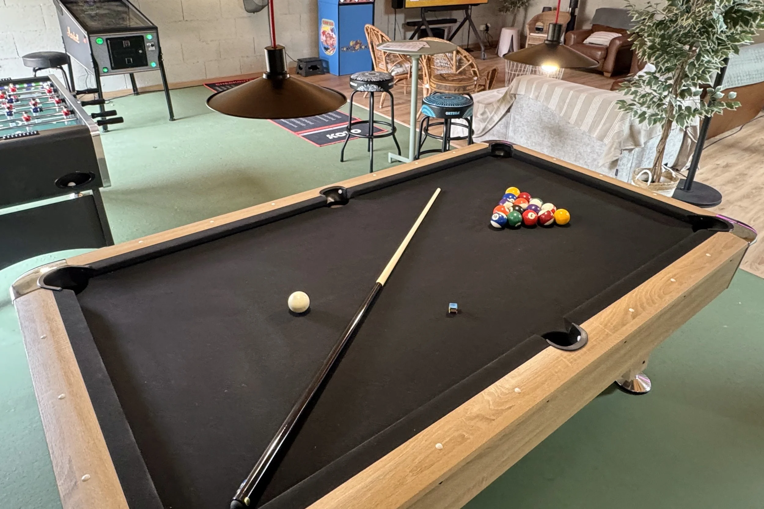 Une table de billard, des billes de billard rassemblées, une queue de billard, une lampe suspendue, un canapé avec une couverture, une plante verte dans un pot en osier, un jukebox bleu, une table avec des chaises en rotin, une aire de jeu de foosball et une salle de détente.