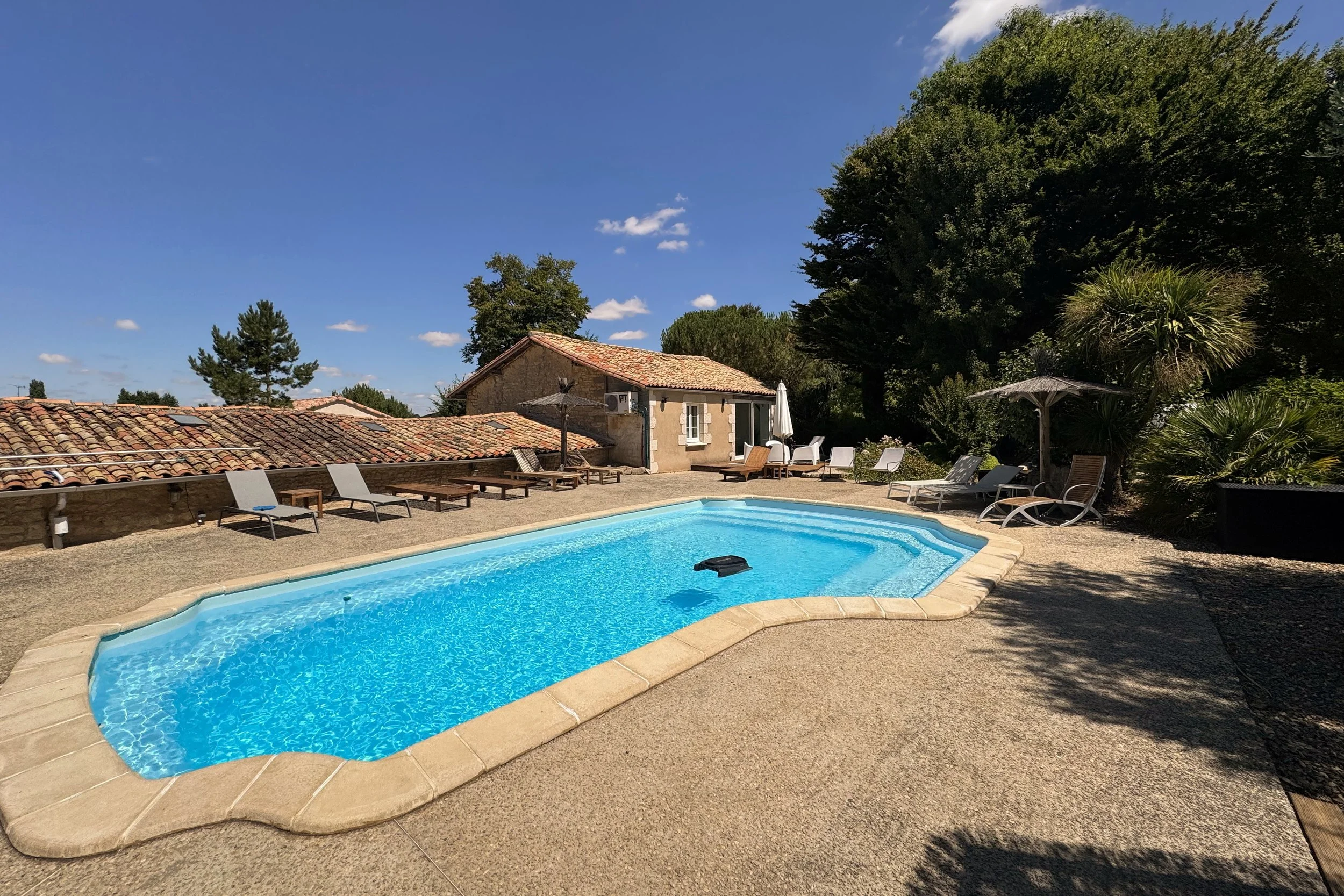 Piscine extérieure dans une cour résidentielle avec des chaises longues, parasols, un bâtiment en pierre, et des grands arbres sous un ciel bleu clair.