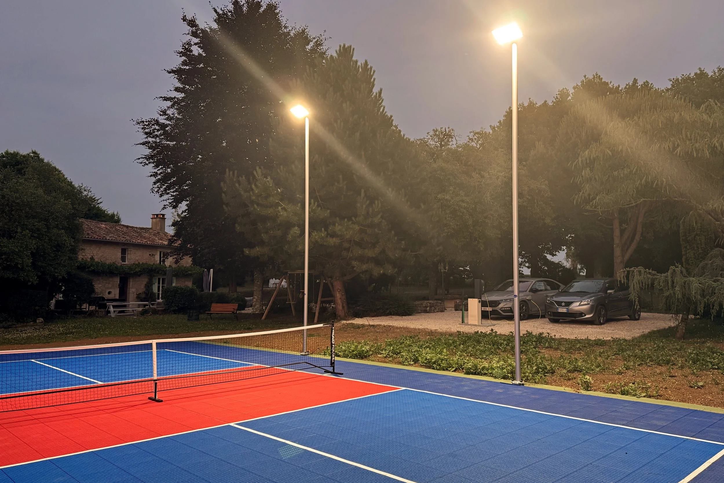 Terrains de tennis extérieurs avec éclairage en soirée, entourés d'arbres et d'une maison, avec des voitures garées en arrière-plan.