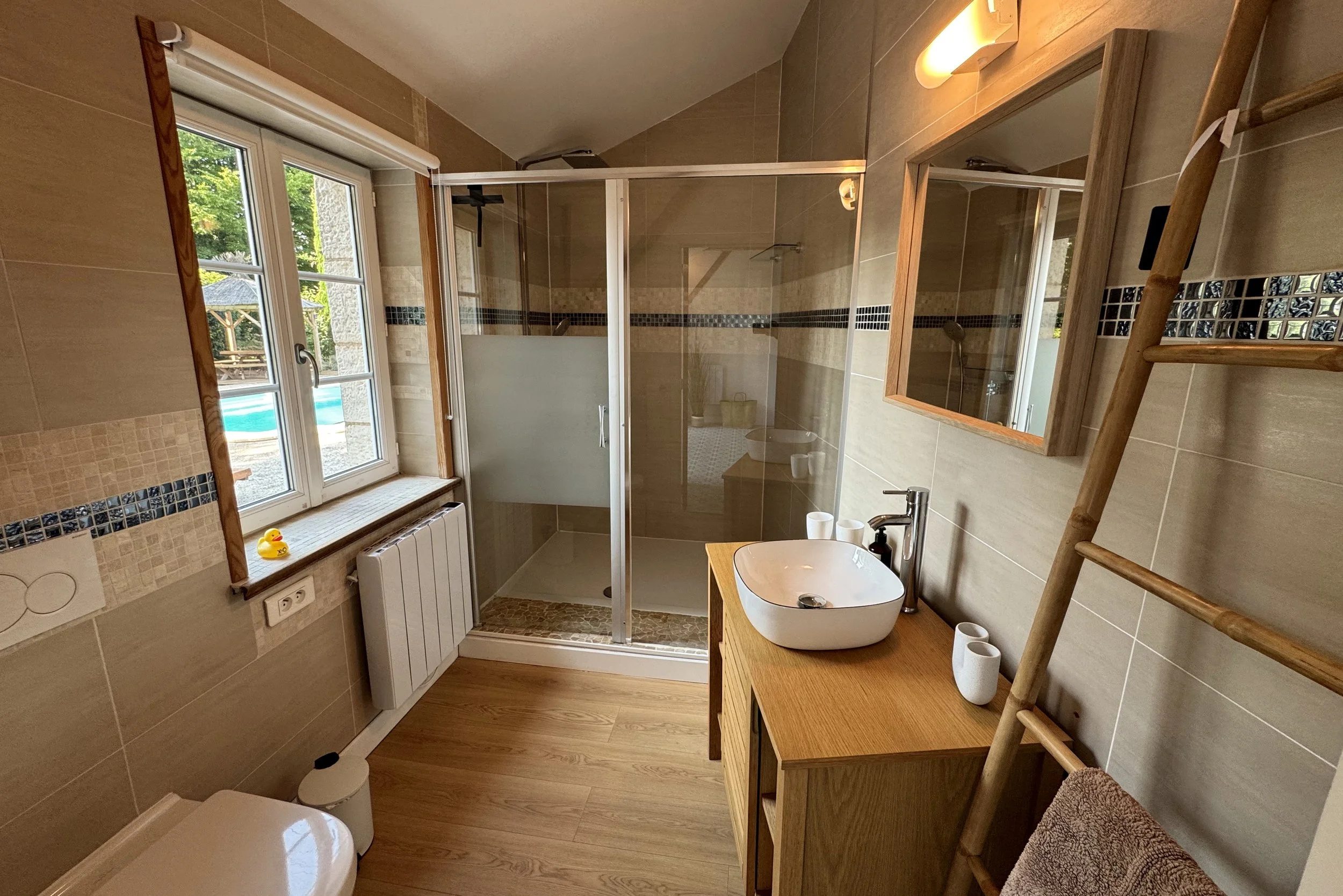 Vue intérieure d'une salle de bain avec une grande fenêtre laissant entrer la lumière naturelle, une cabine de douche en verre avec des parois en aluminium, un lavabo rond posé sur une console en bois, un miroir, un radiateur électrique blanc, une échelle en bois pour suspendre des serviettes, et divers objets de toilette.
