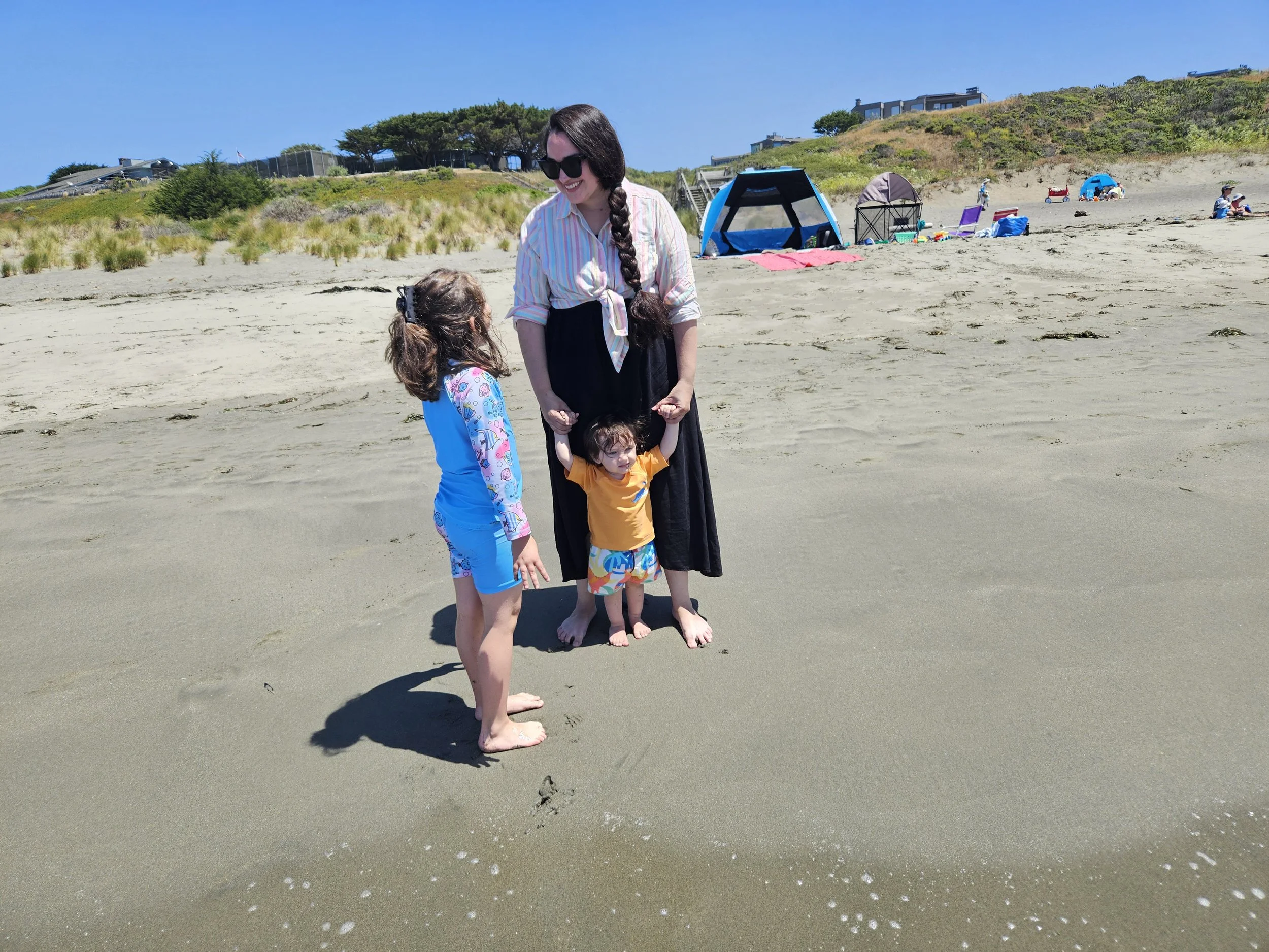A woman with long braided dark hair wearing sunglasses, a light striped shirt, and a black skirt, holding hands with a small girl in colorful swimwear on a sandy beach.