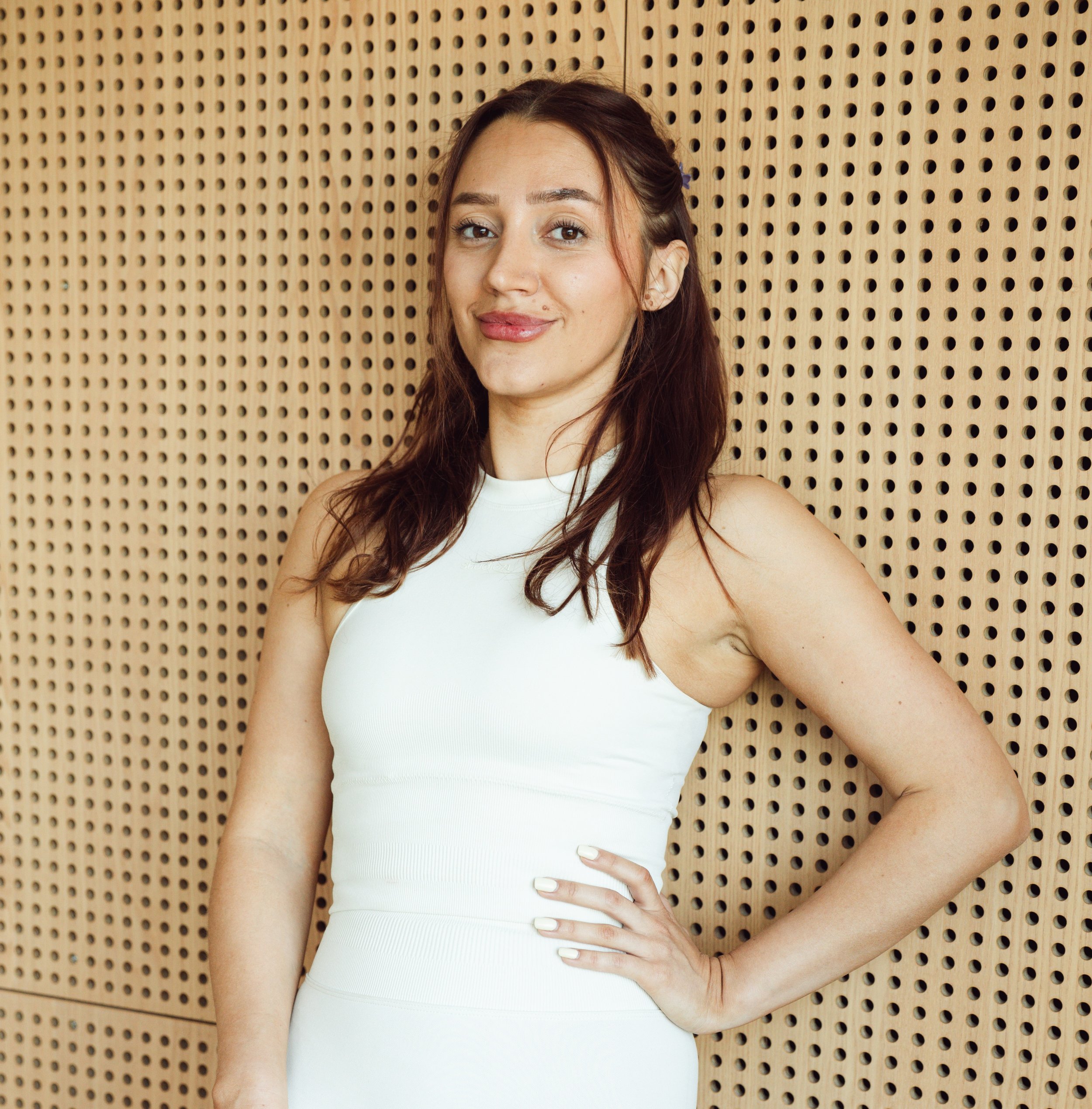 A woman with long brown hair wearing a white sleeveless top, standing in front of a perforated wooden wall, with one hand on her waist and smiling at the camera.