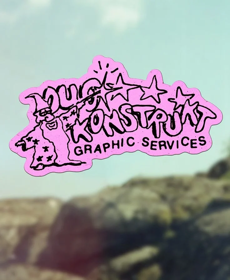 A pink die-cut sticker with black handwritten-style text that reads "DUNS TASTY GRAPHIC SERVICES" and includes a cartoon witch and stars, placed on a transparent surface with a blurred outdoor background.