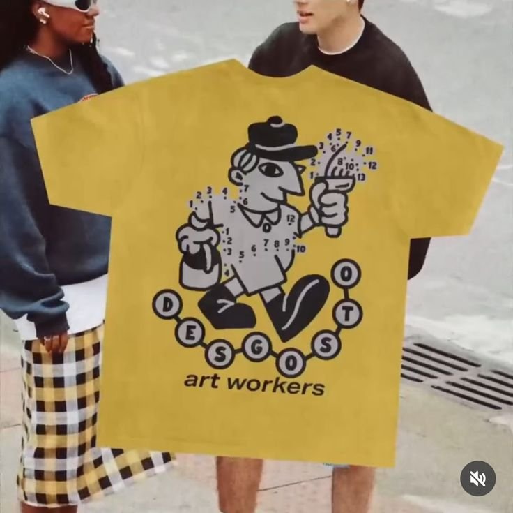 Two people holding a yellow giant T-shirt with a cartoon figure of a man, along with chemical element symbols and the words "art workers" printed on it.